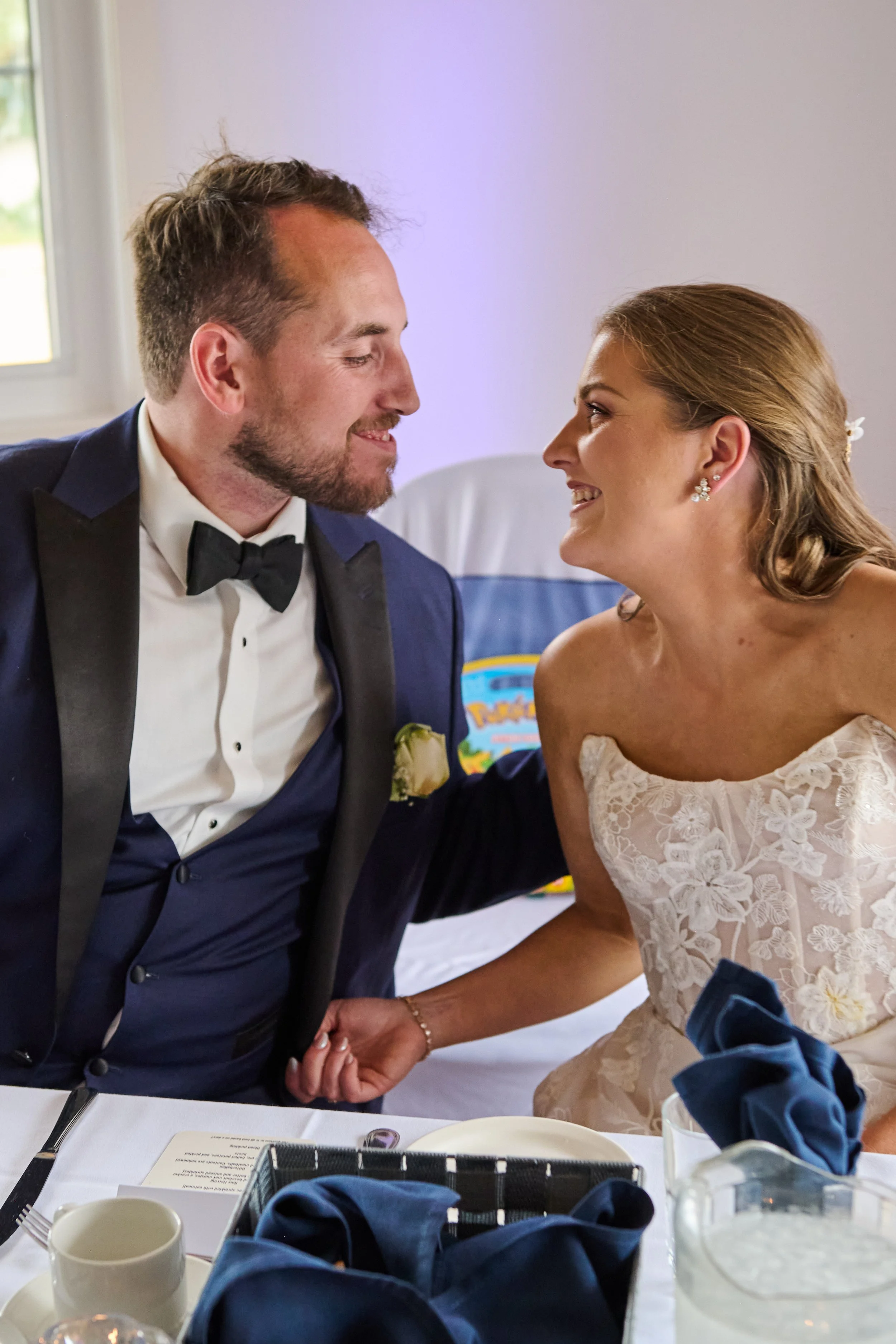 A groom and bride smiling at each other during a wedding reception, sitting at a table with navy blue napkins, a plate, and a glass.