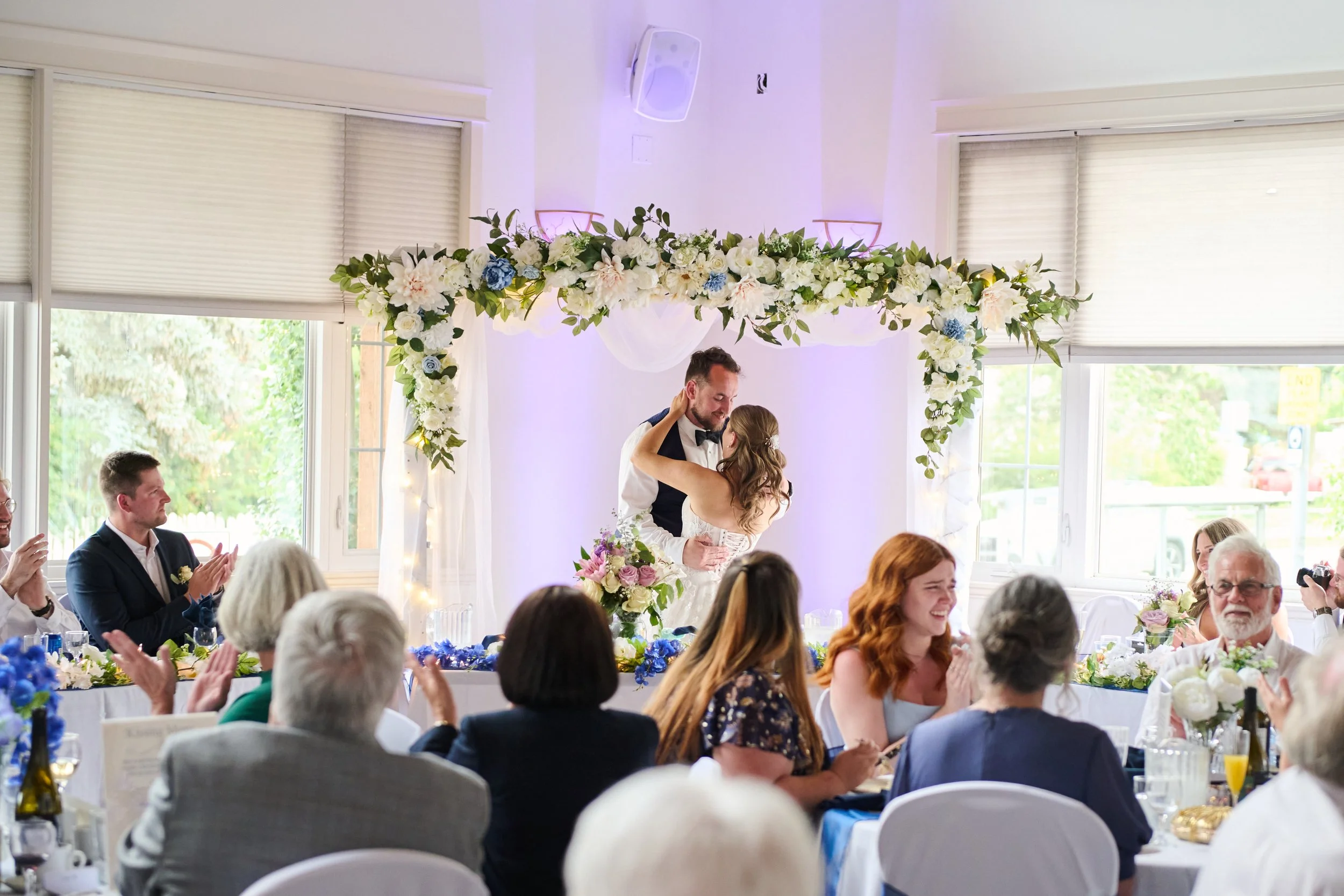 Bride and groom sharing a dance at their wedding reception under a floral arch, surrounded by seated guests clapping and smiling.