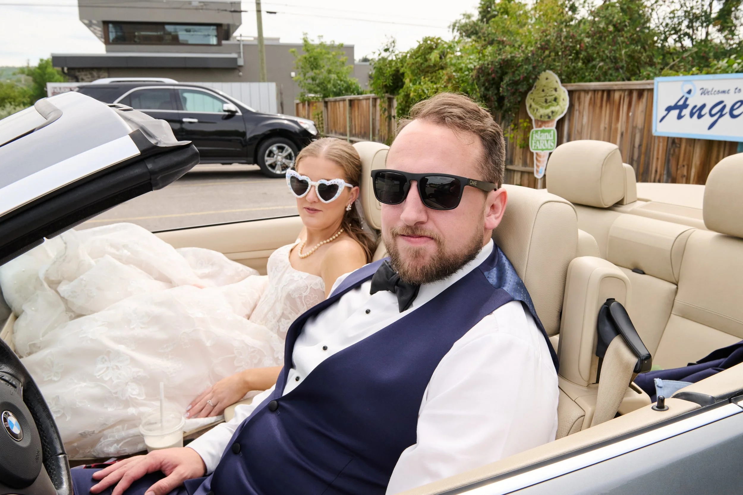 A man and a woman dressed in wedding attire sitting in a convertible car, both wearing sunglasses. The woman is in a wedding gown with heart-shaped sunglasses, and the man is in a tuxedo with dark sunglasses, holding a drink with a straw.