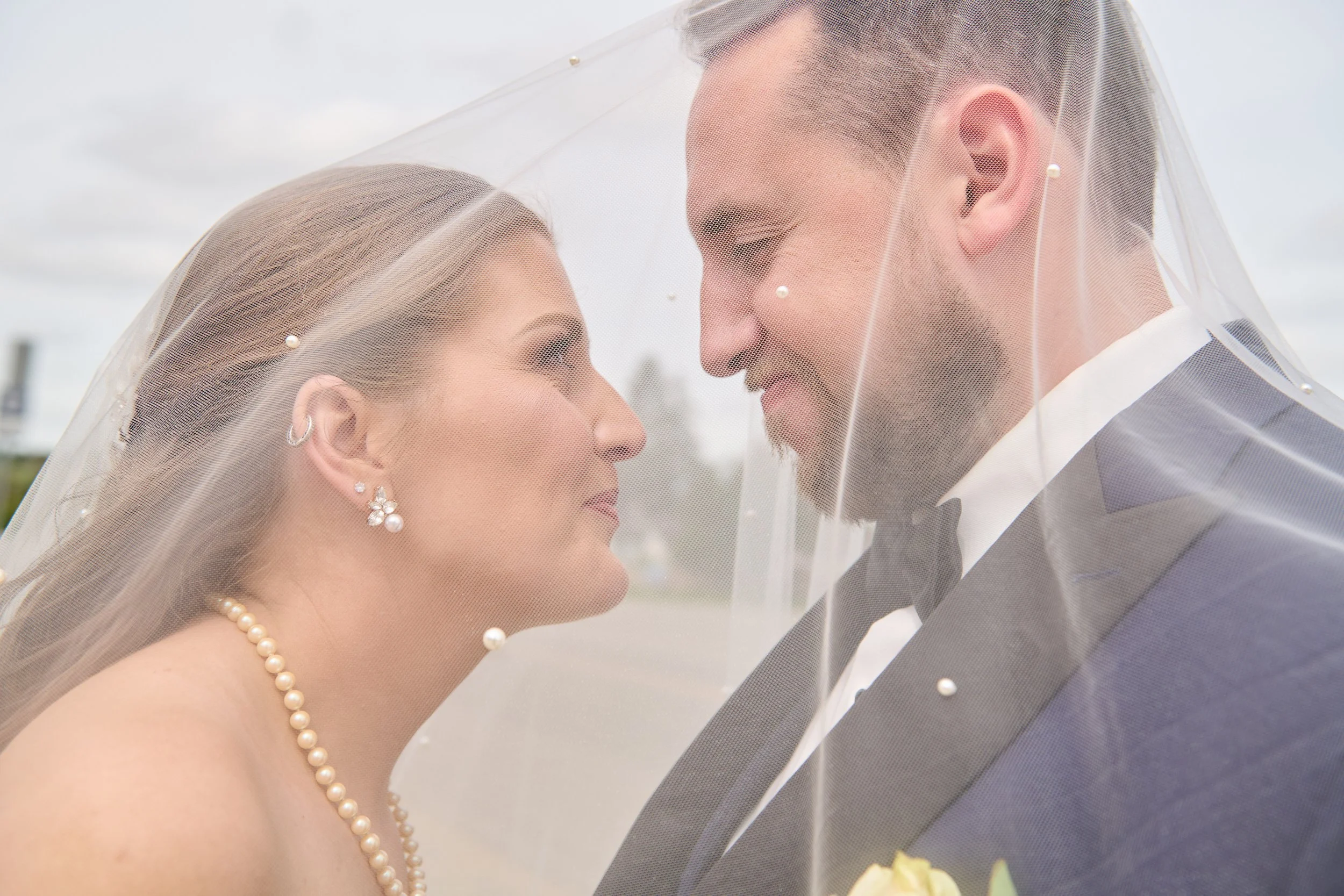 A bride and groom face each other under a veil, smiling softly during their wedding.