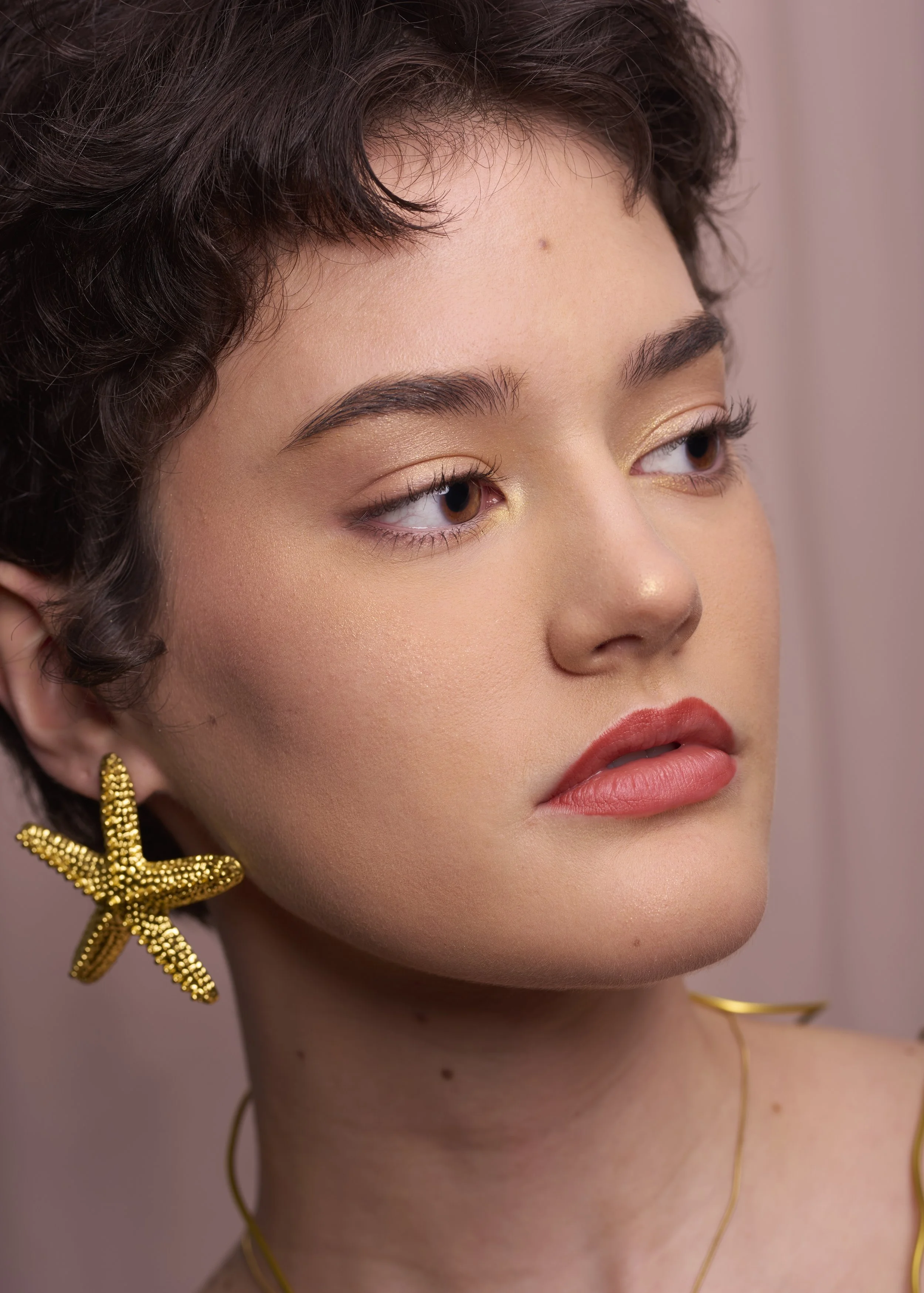 Close-up of a woman with short dark hair, wearing gold star-shaped earrings and a delicate gold necklace, with natural makeup and a soft expression.