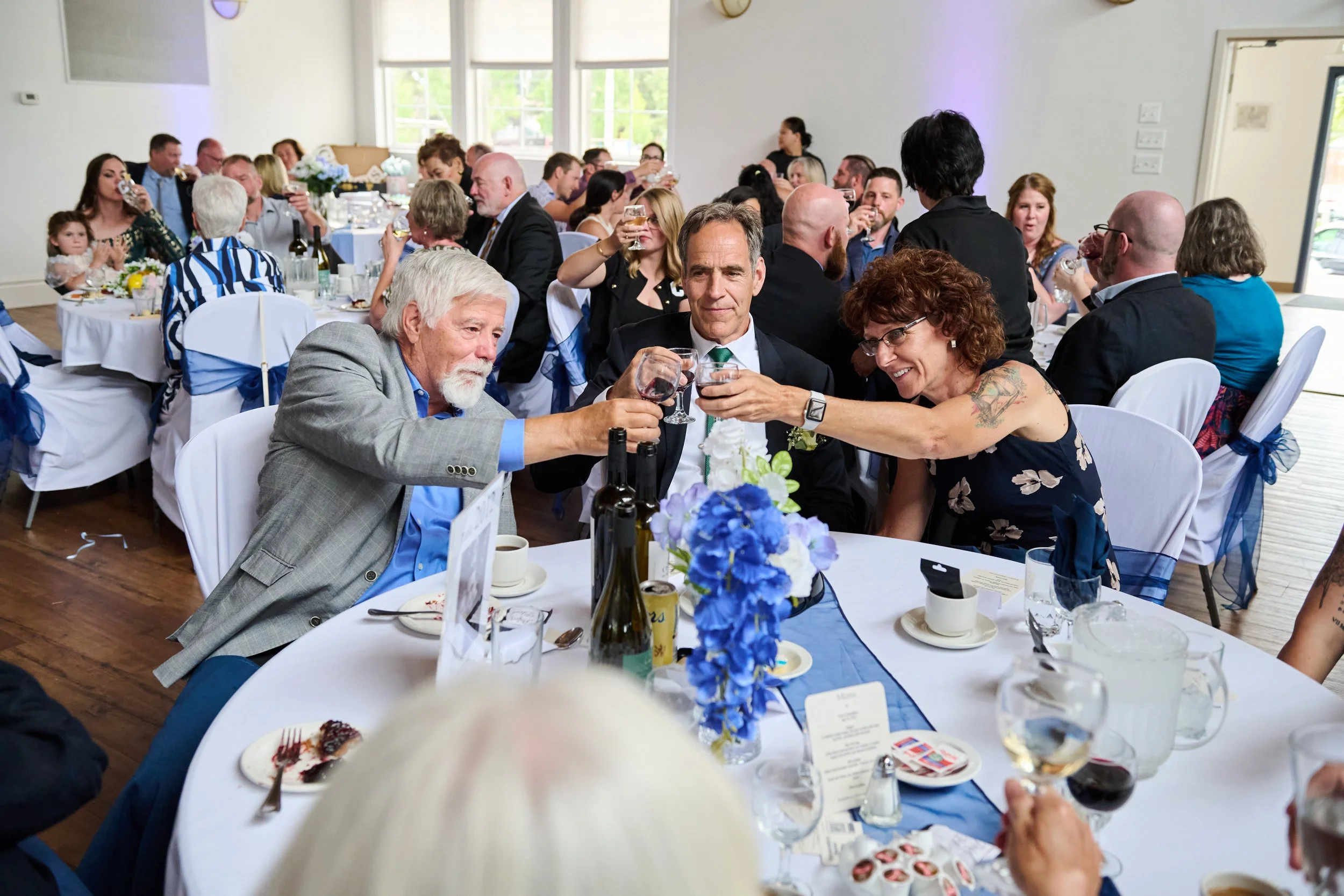 People at a banquet table toasting with glasses of wine at a wedding reception or formal event, decorated with blue accents and flowers.