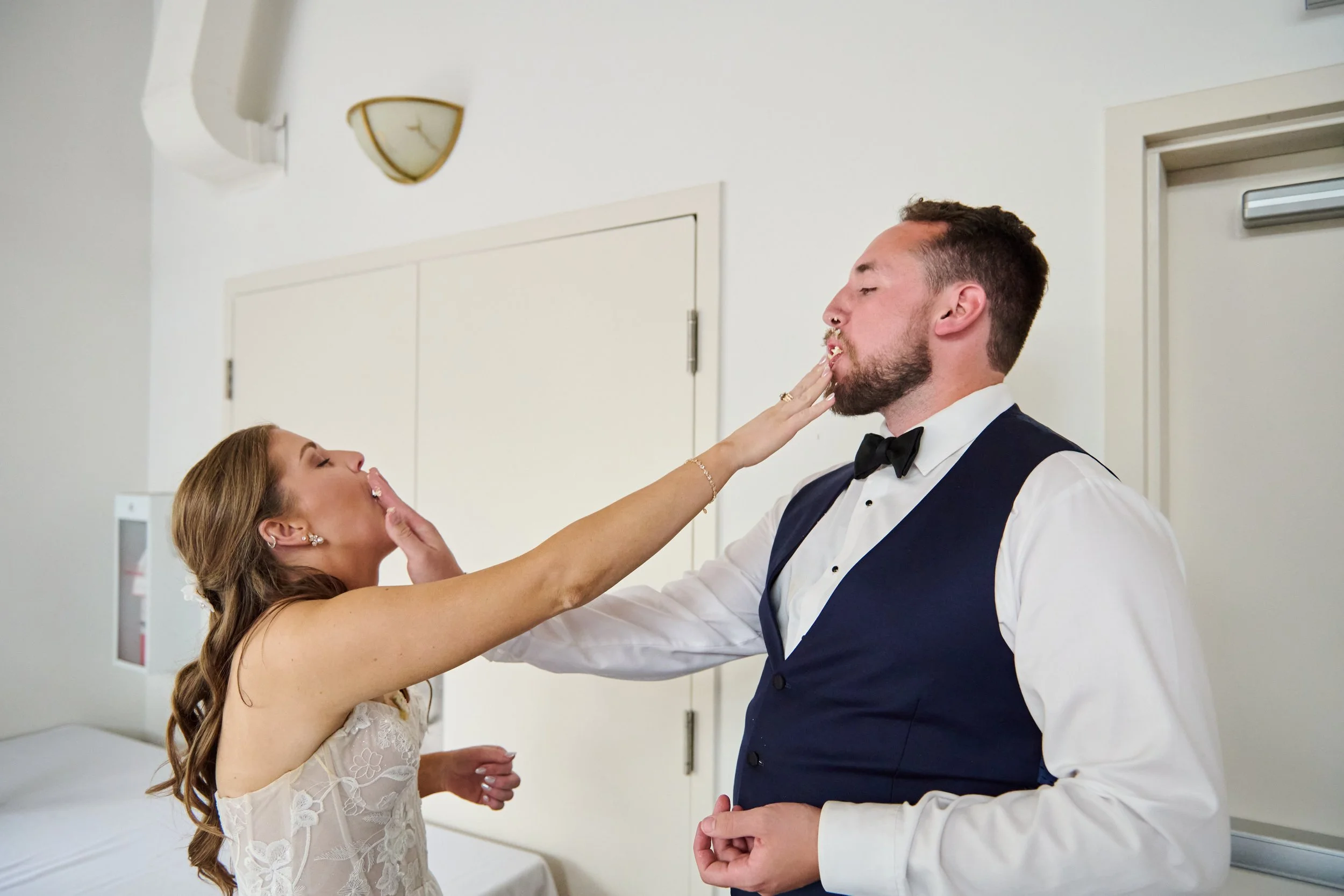 A bride and groom are playfully slapping each other's faces during their wedding, inside a room with white walls and closed double doors.
