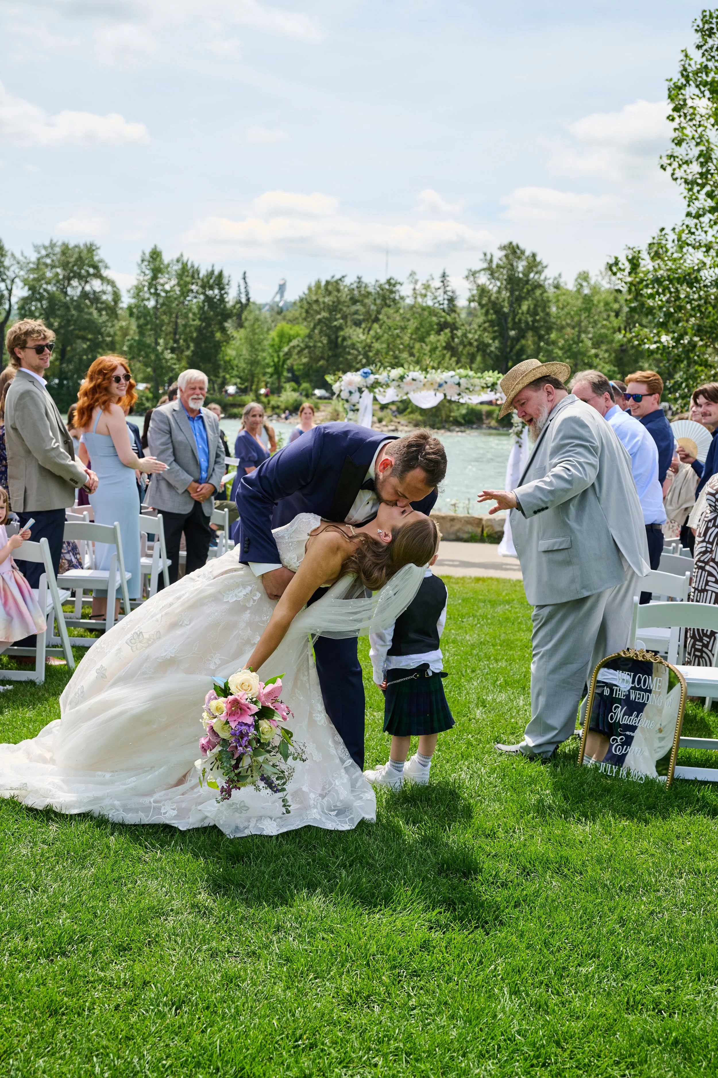 A wedding ceremony taking place outdoors by a river on a sunny day, with the bride and groom kissing and a small child in a plaid skirt standing between them, while guests watch.