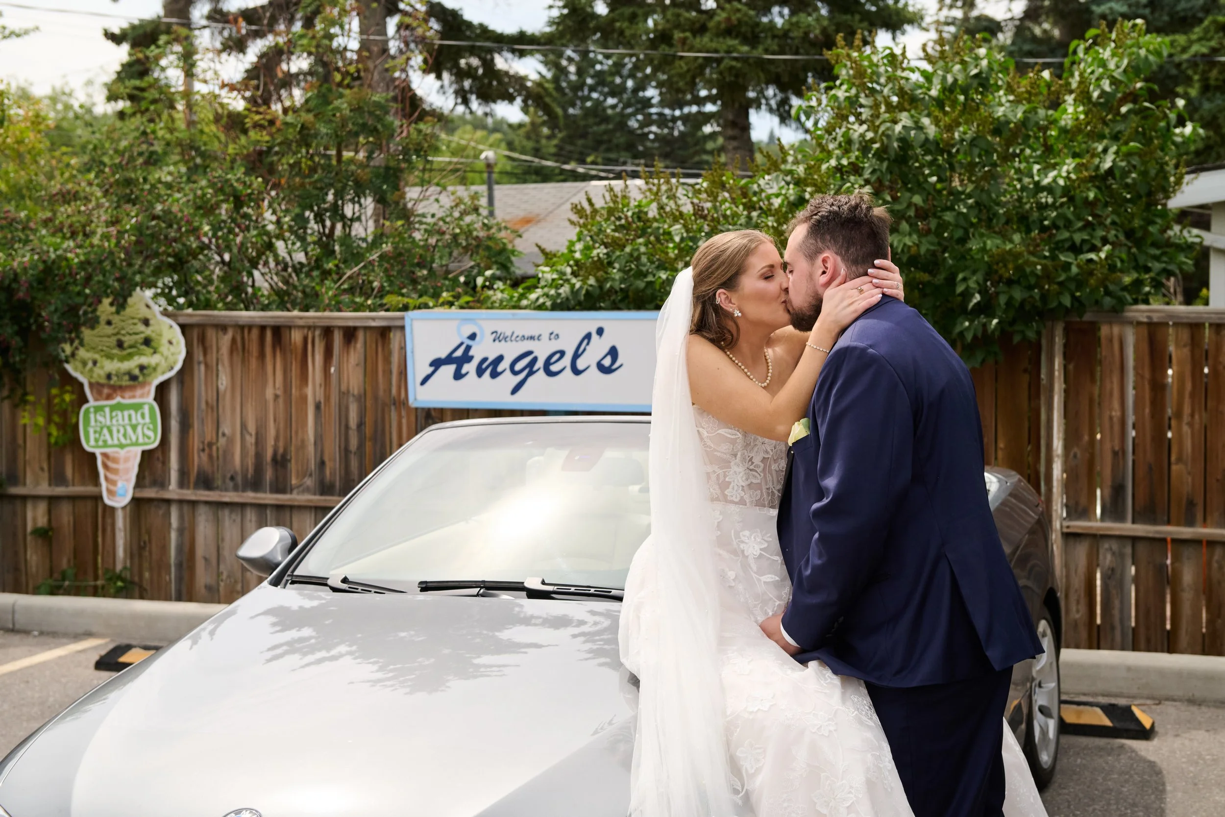 A bride and groom sharing a kiss on a silver car in a parking lot, with a sign reading 'Welcome to Angel's' and a wooden fence with trees in the background.