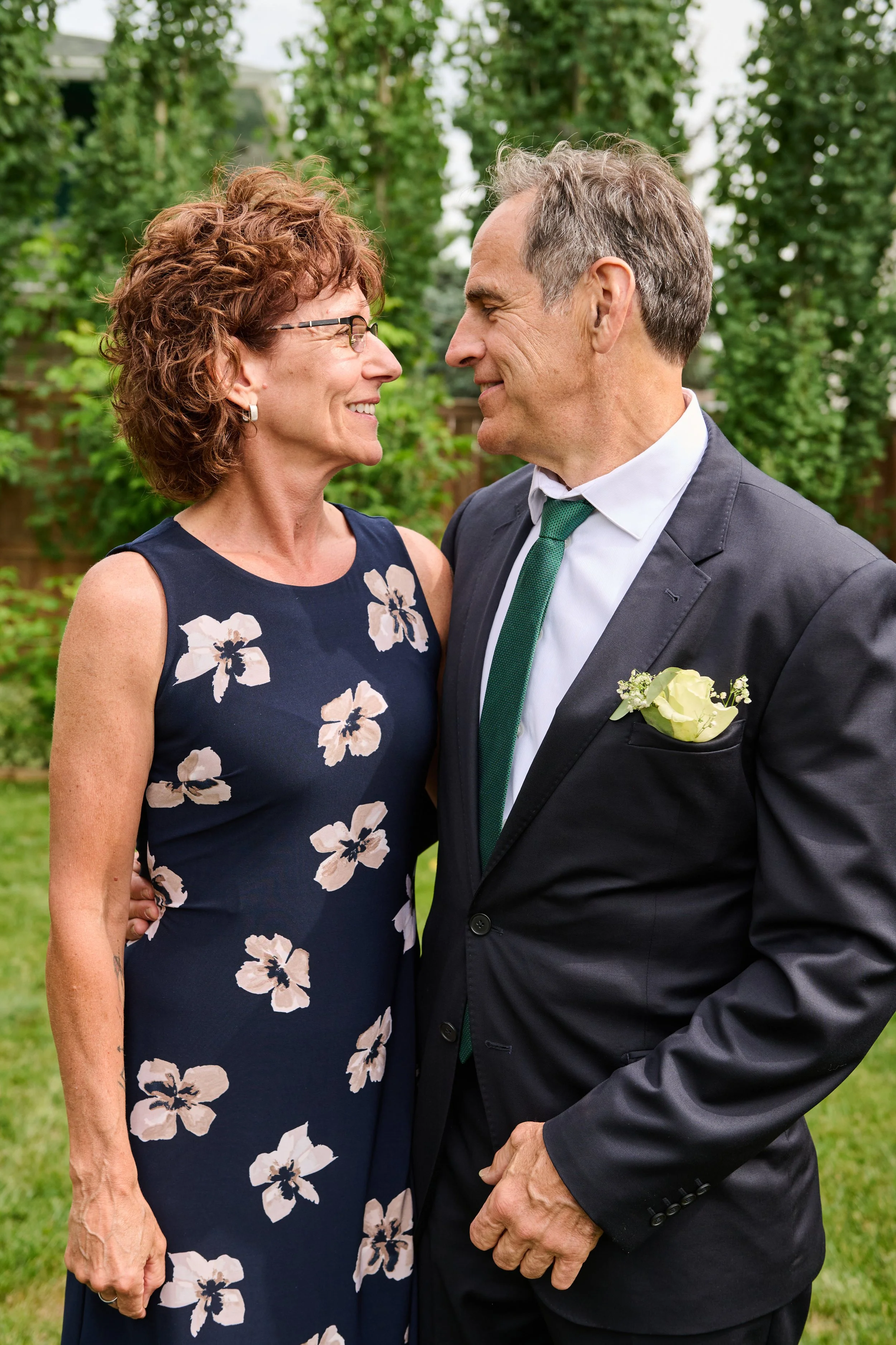 A middle-aged woman and man are standing outdoors, looking into each other's eyes and smiling. The woman is wearing a sleeveless navy dress with a floral pattern, and the man is wearing a dark suit with a white shirt, green tie, and a white flower bo