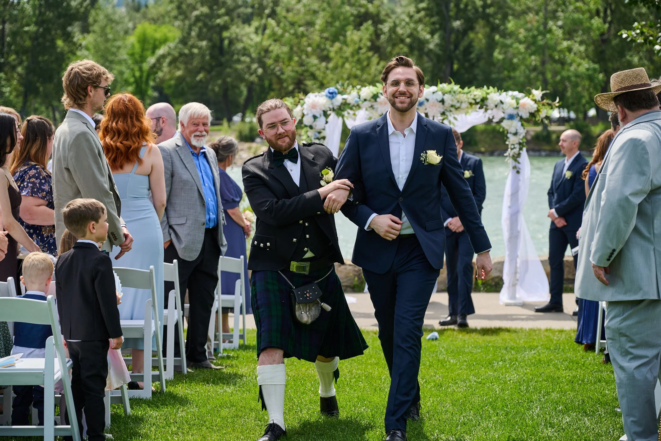 A wedding ceremony outdoors with two men, one in a kilt and the other in a navy suit, walking down the aisle together, surrounded by guests and a floral arch.