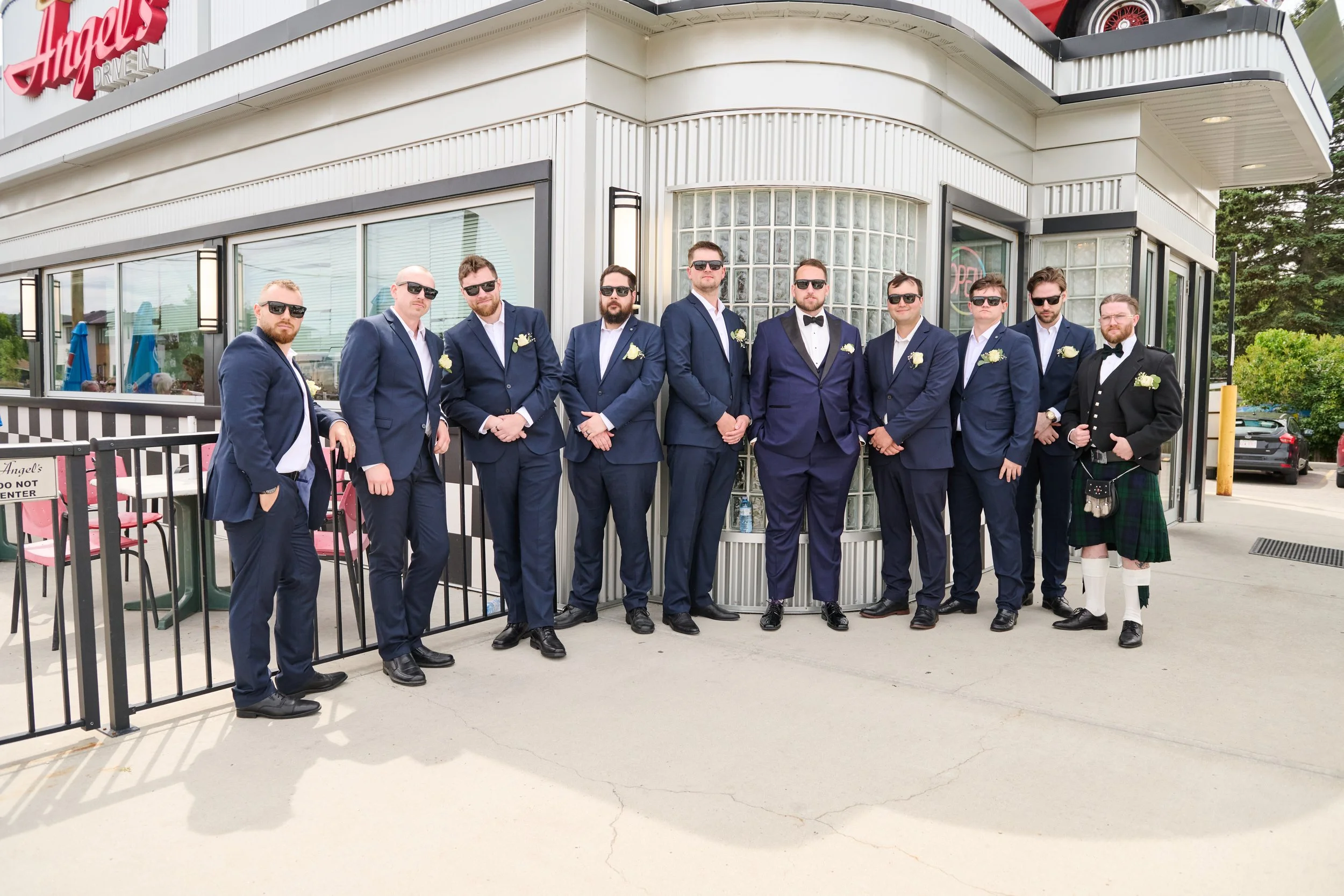 A group of eleven men in formal suits, most with sunglasses, standing outside a diner with a red sign that says 'Angel's'. One man on the far right wears a traditional Scottish kilt and jacket. They are posing for a photo, likely at a wedding or spec