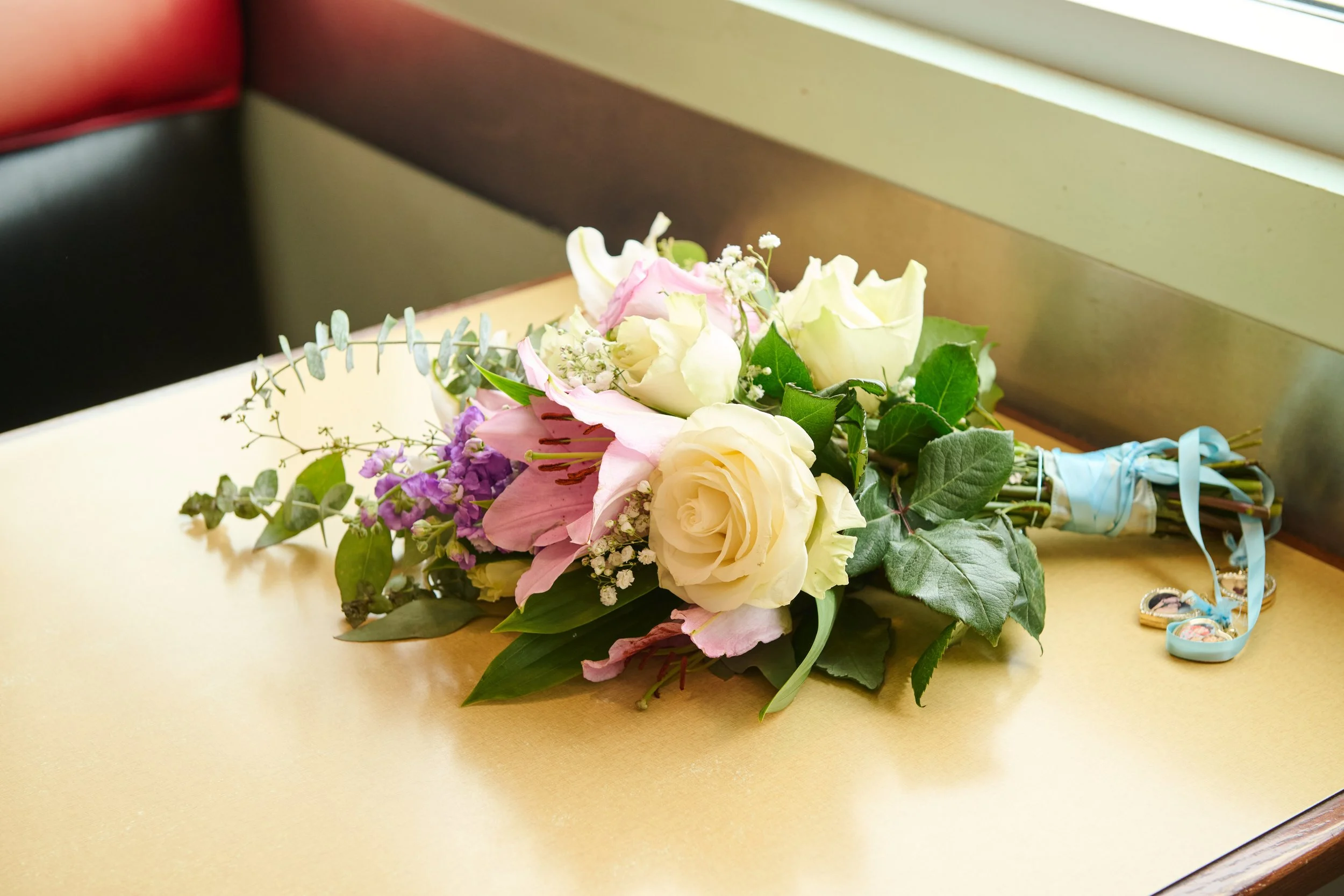 A bouquet of pink and white flowers with greenery, tied with light blue ribbons, placed on a yellow table near a window.