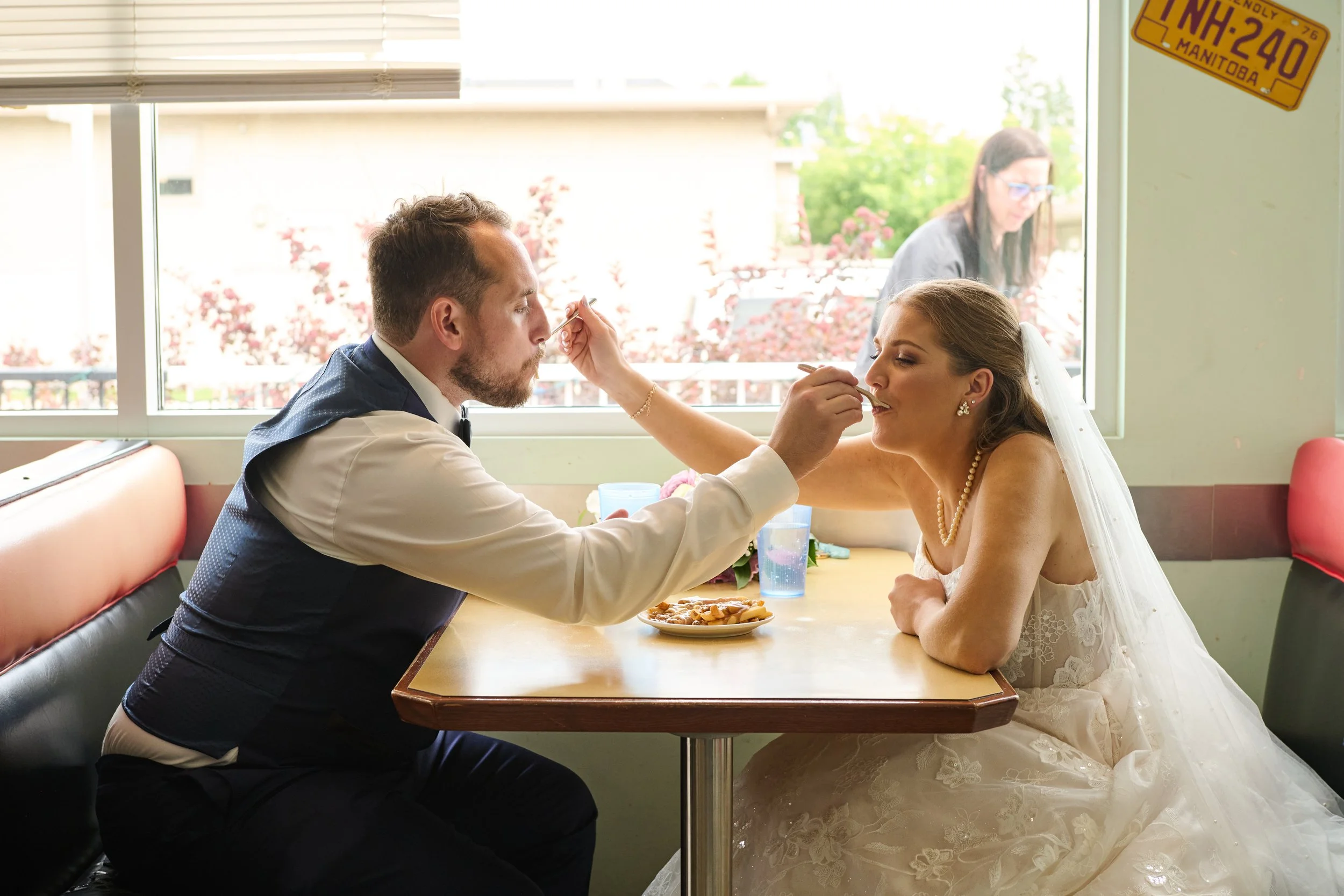 A bride and groom sitting at a restaurant table with the groom feeding the bride. The bride is wearing a wedding dress and the groom is dressed in a vest and bow tie, with a window and a woman outside in the background.