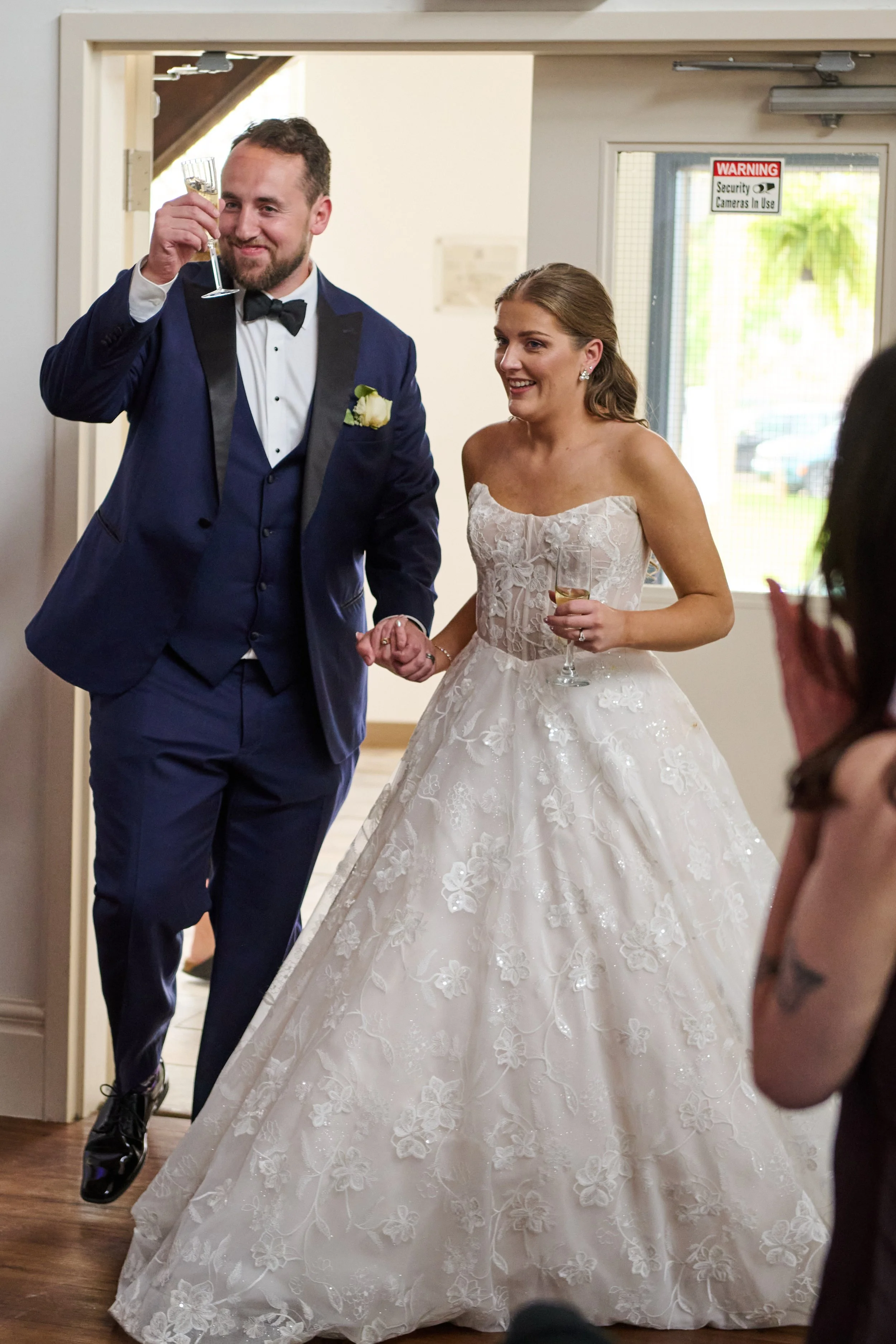A man in a blue tuxedo with a bow tie and boutonniere is holding a glass of champagne and smiling as he leads a woman in a wedding dress holding a glass of champagne at a wedding reception.