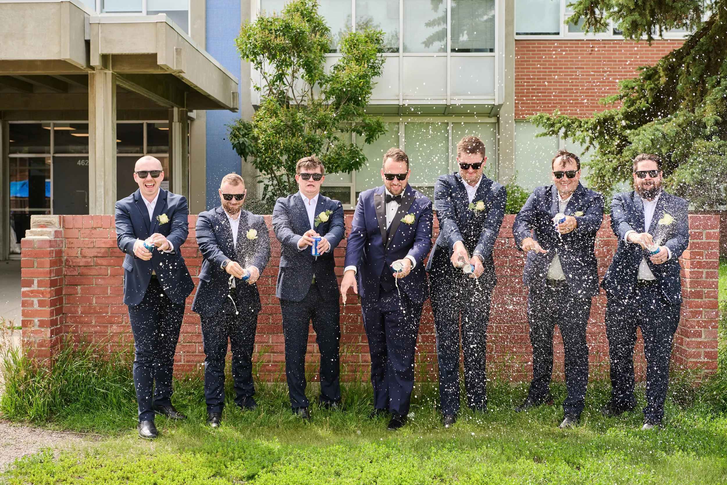 Group of eight men in suits celebrating outside, spraying champagne, standing in front of a brick wall with trees and buildings in the background.