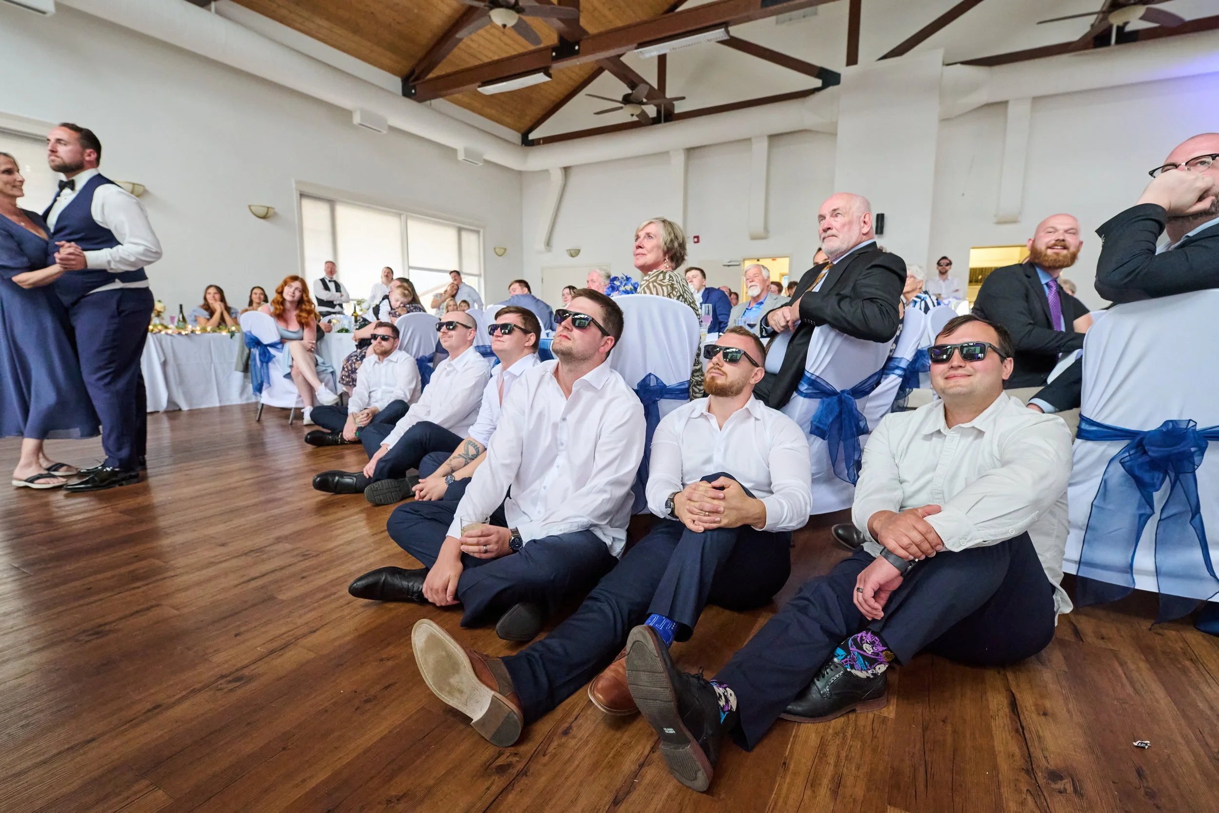Attendees at a wedding reception, with some sitting on the floor wearing sunglasses, others sitting at tables in decorated chairs, and a couple dancing in the background.