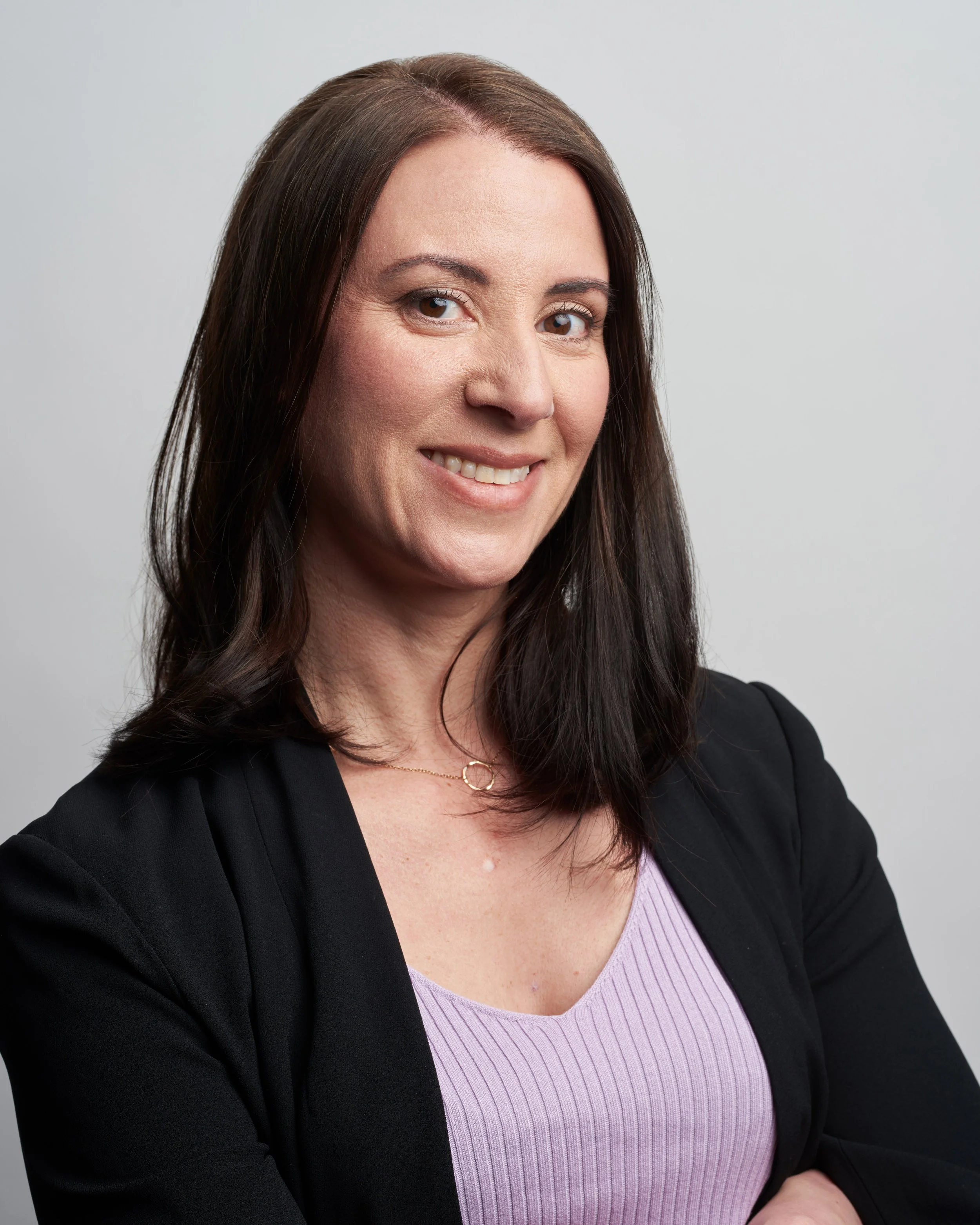 Woman smiling, wearing a black blazer and lavender top, neutral background.