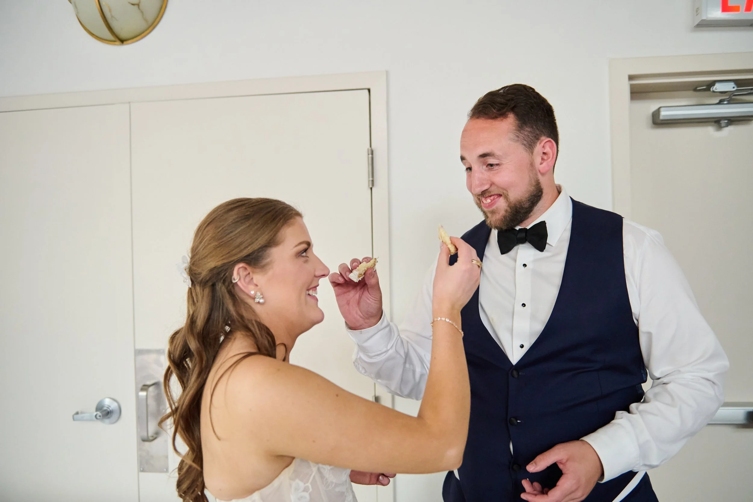 A bride and groom sharing a playful moment, with the bride feeding the groom a piece of cake, both smiling and dressed in wedding attire.