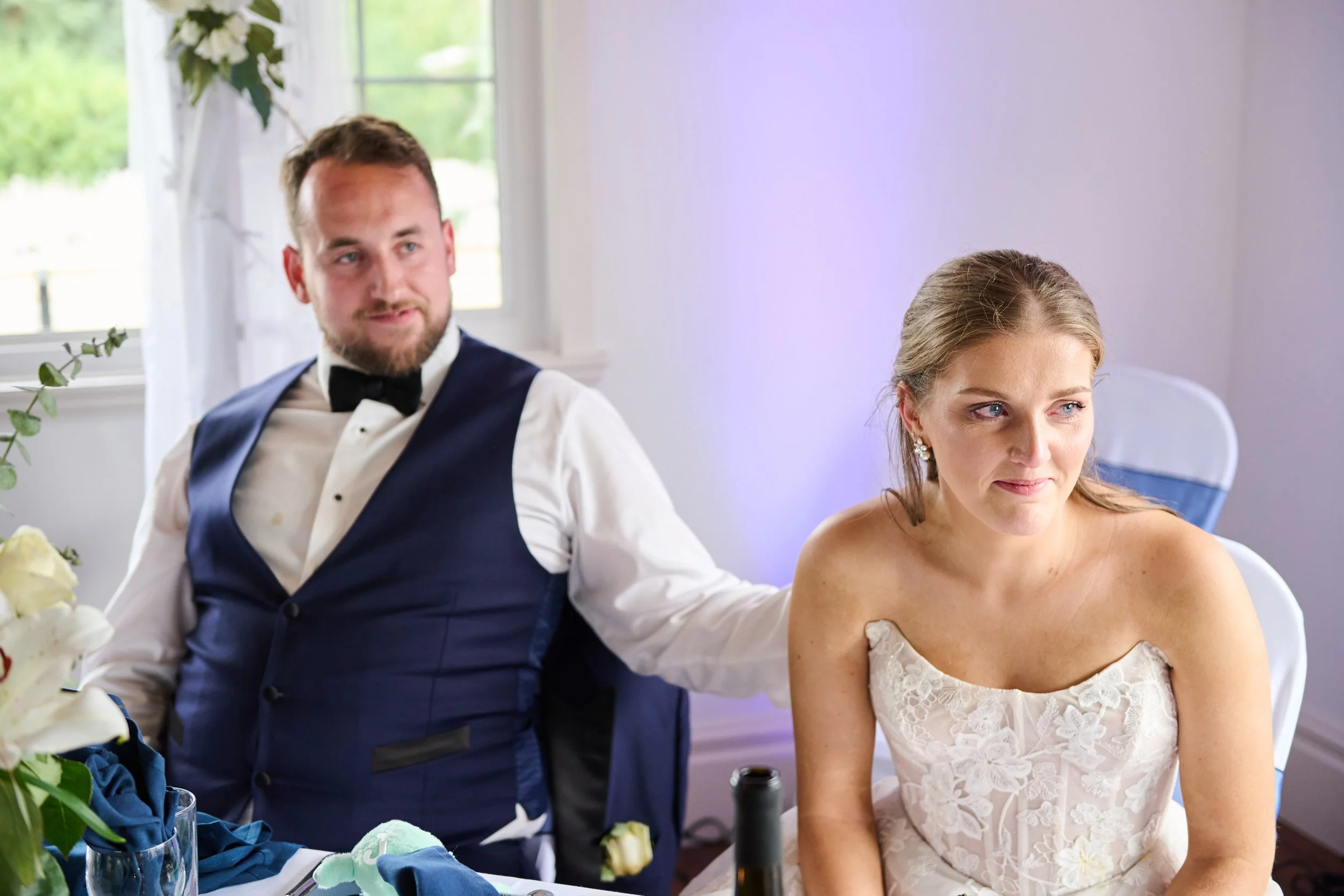 A groom and bride sitting next to each other at a wedding reception. The groom is wearing a tuxedo with a bow tie, and the bride is wearing a strapless wedding dress. They are inside a decorated room with flowers and a window in the background.