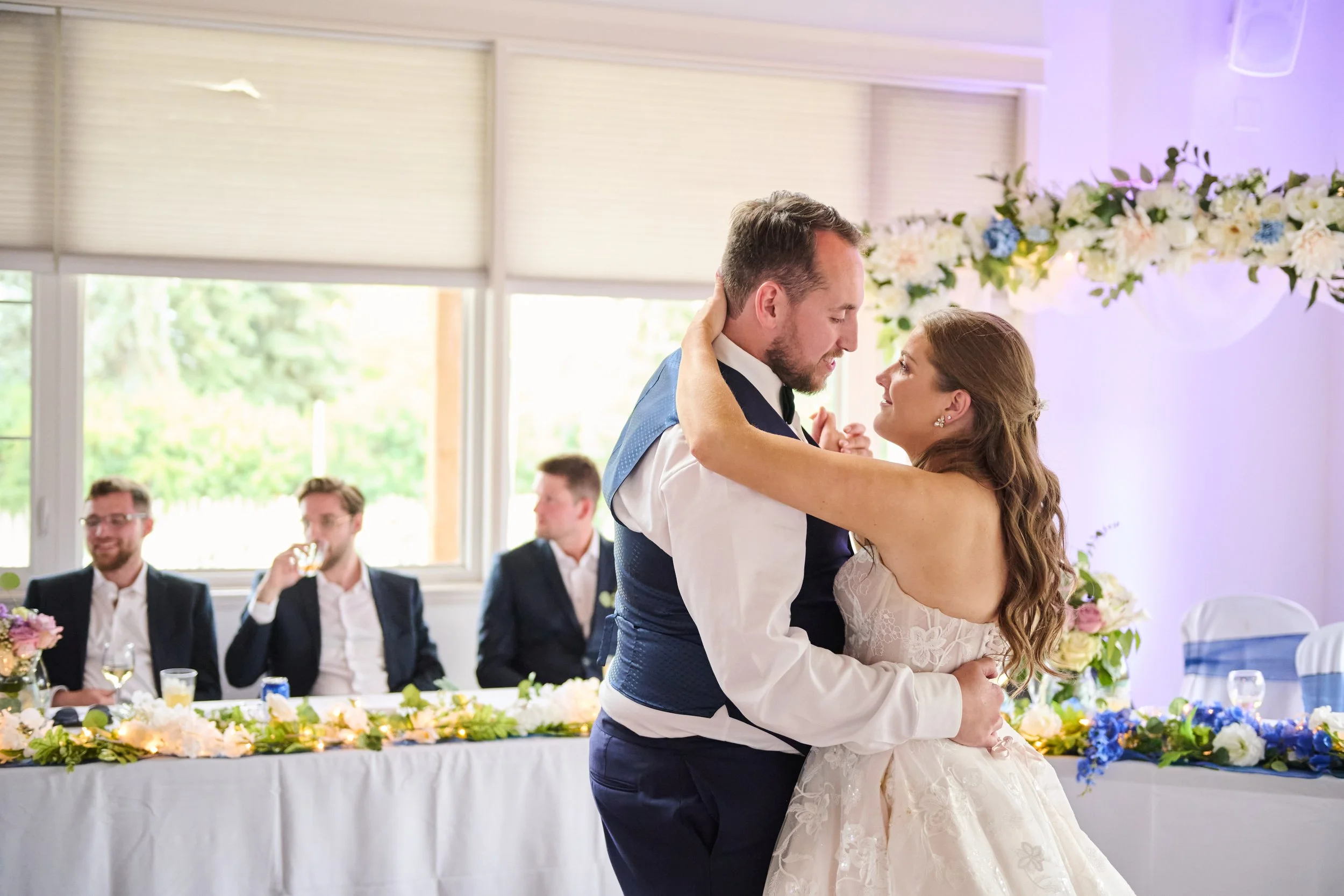 Bride and groom sharing their first dance at a wedding reception with seated guests in the background, decorated with flowers.