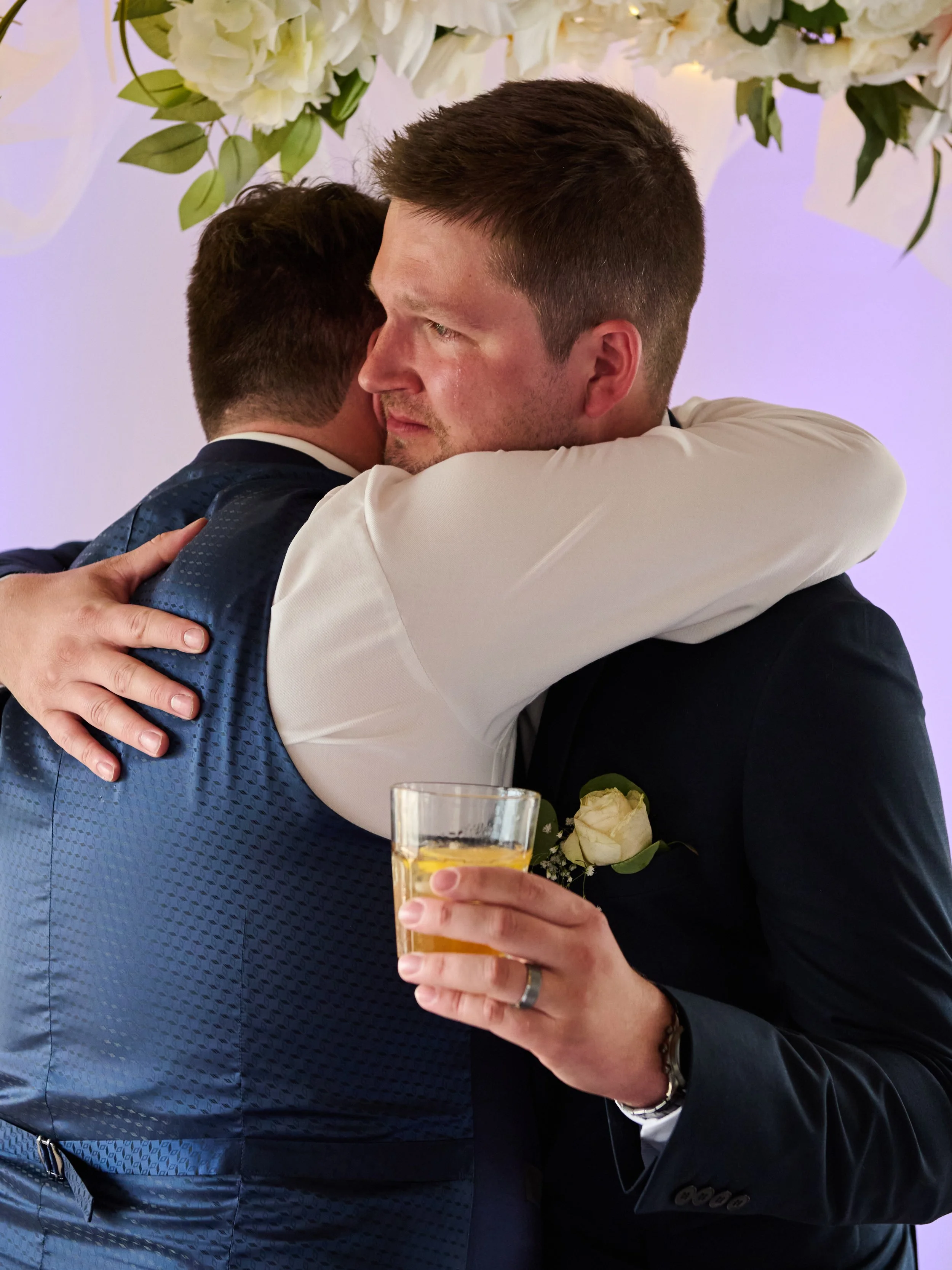 Two men embracing at a celebration, one holding a drink, surrounded by white flowers.
