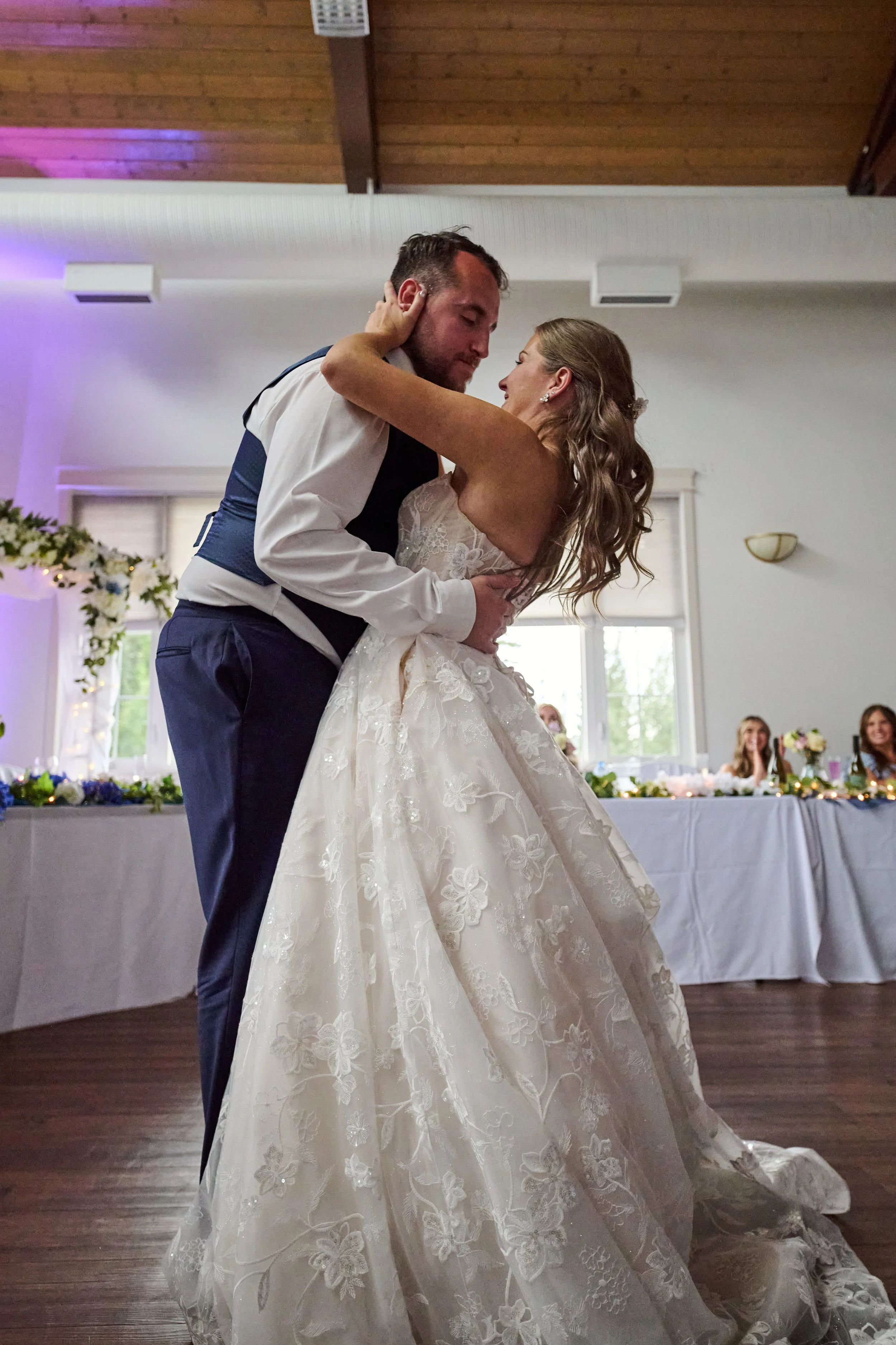 Bride and groom dancing at a wedding reception, with the bride in a white lace wedding gown and the groom in formal attire, inside a decorated hall with large windows and a table with guests in the background.