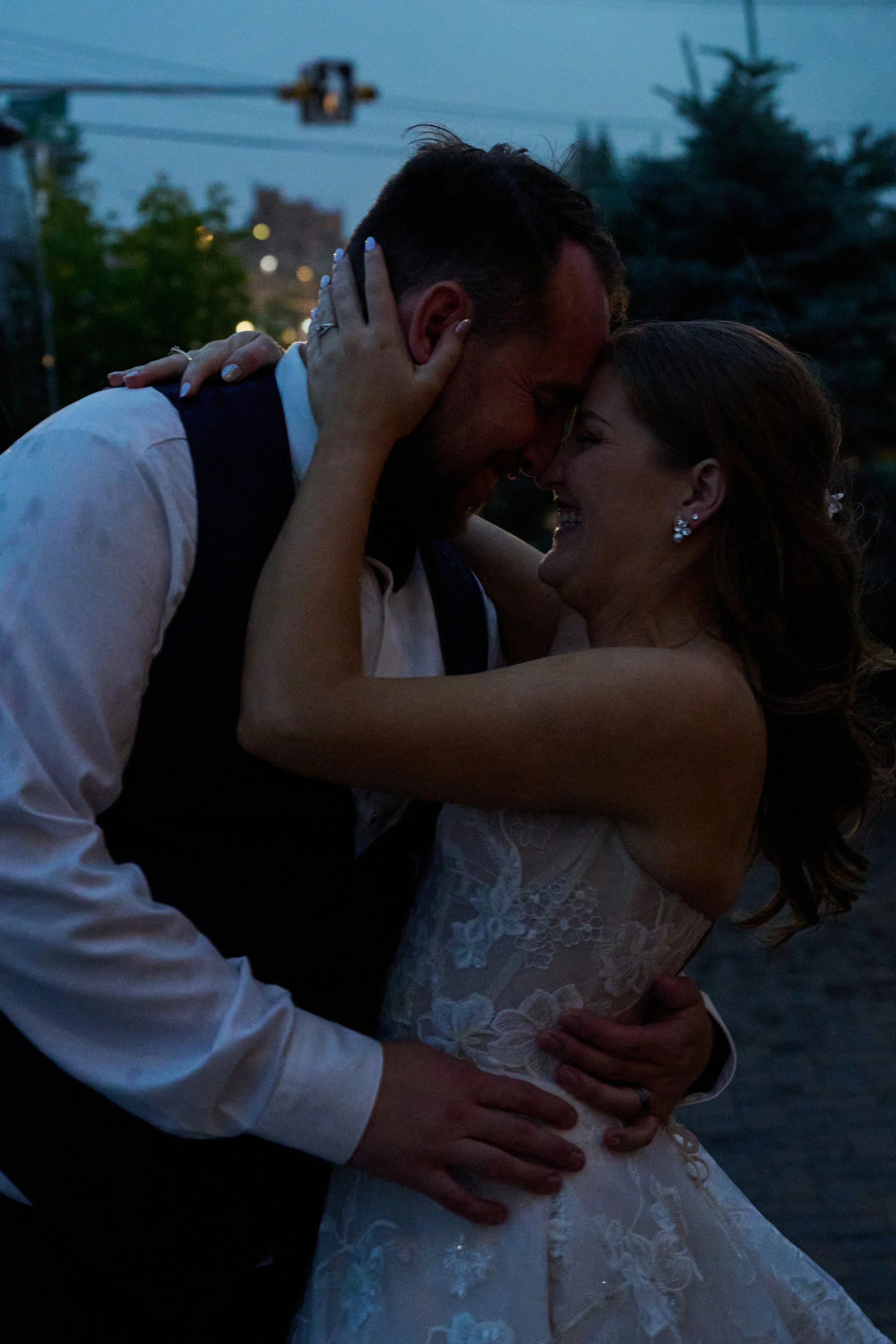 A couple in wedding attire embracing and smiling outdoors at dusk.