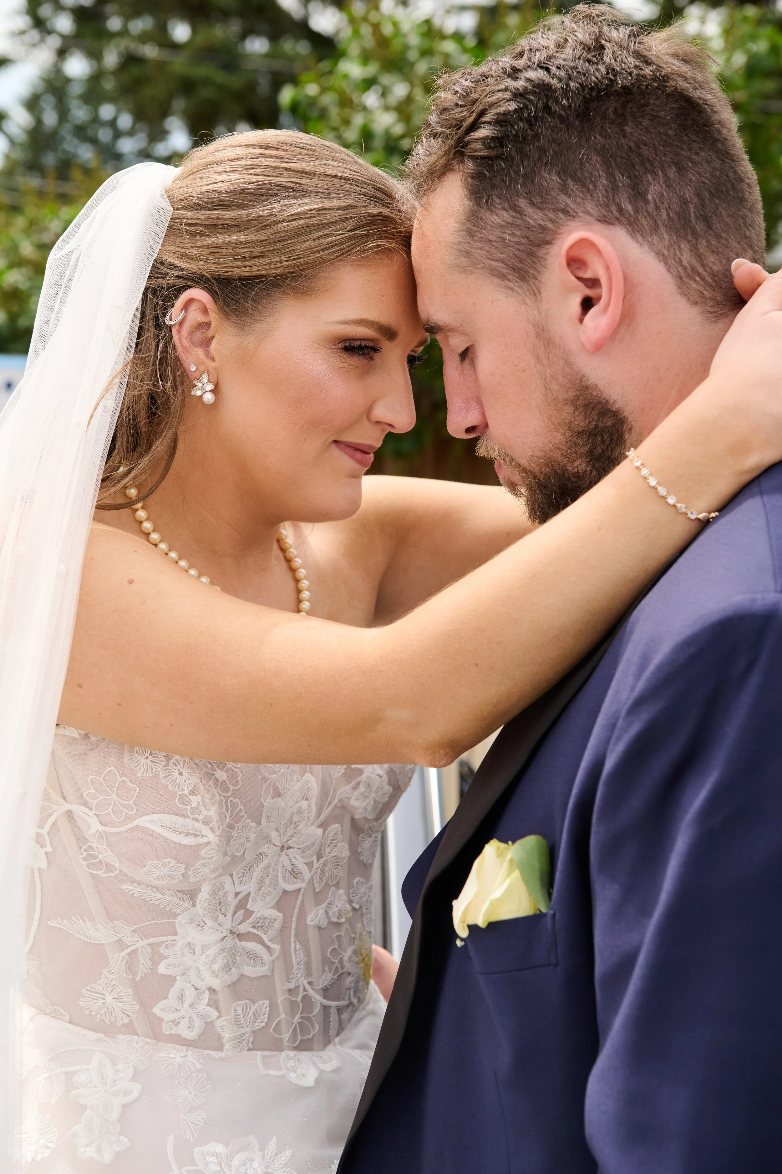 A newlywed couple with their foreheads touching, eyes closed, during their wedding ceremony outdoors. The bride is wearing a lace wedding dress, pearl necklace, earrings, and a veil. The groom is in a dark suit with a boutonniere.
