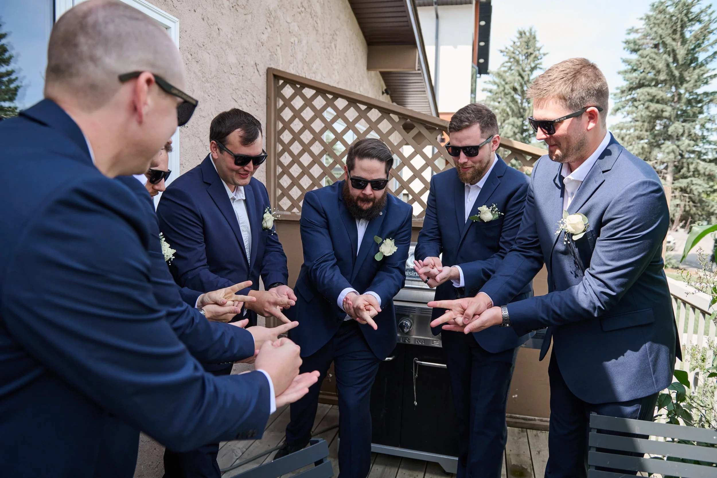 A group of six men in matching navy suits and sunglasses standing on a balcony, with some wearing white boutonnieres, all pointing at something and engaged in a conversation, with trees in the background.