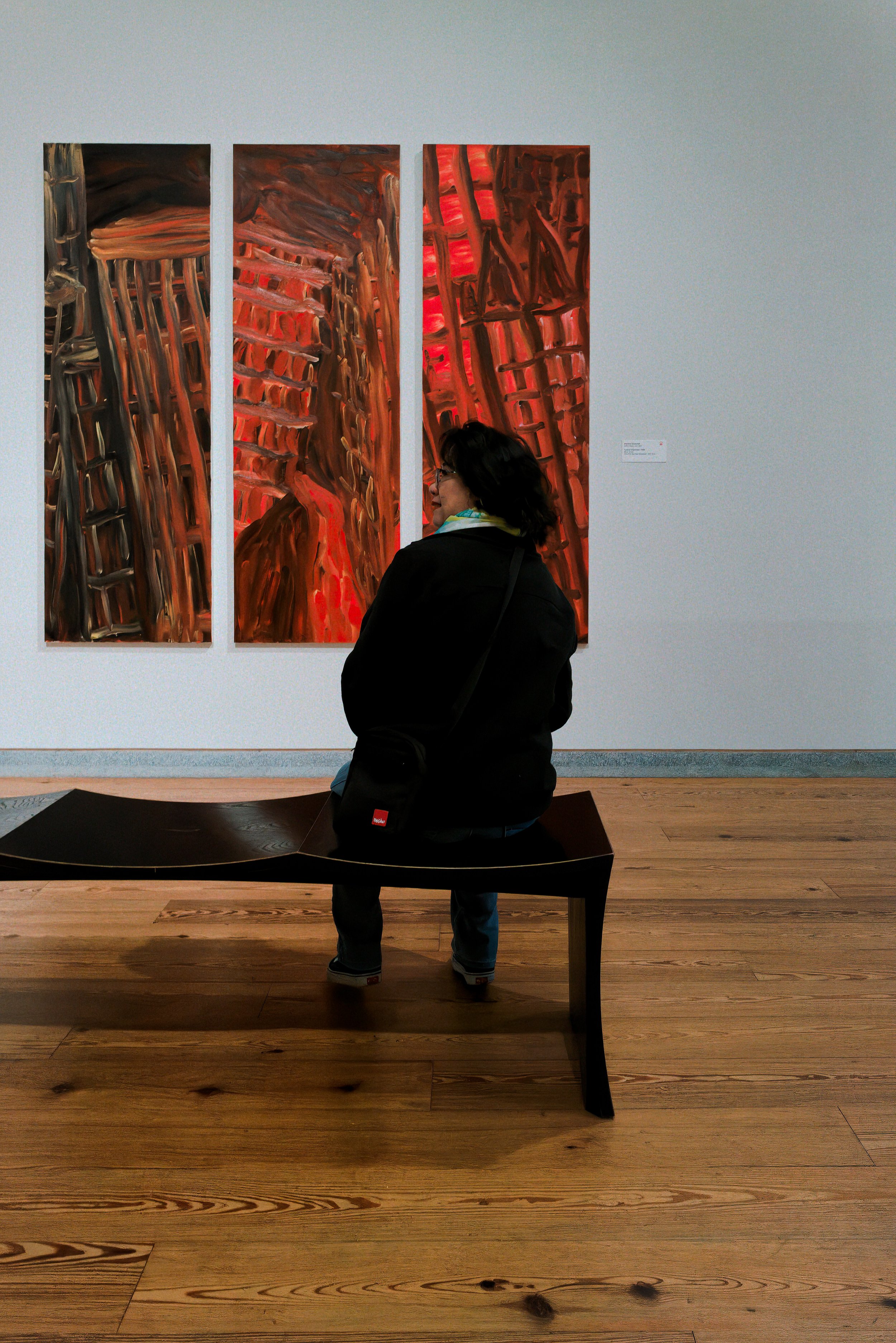 Person seated on a museum bench looking at three red and brown abstract paintings on a gallery wall.