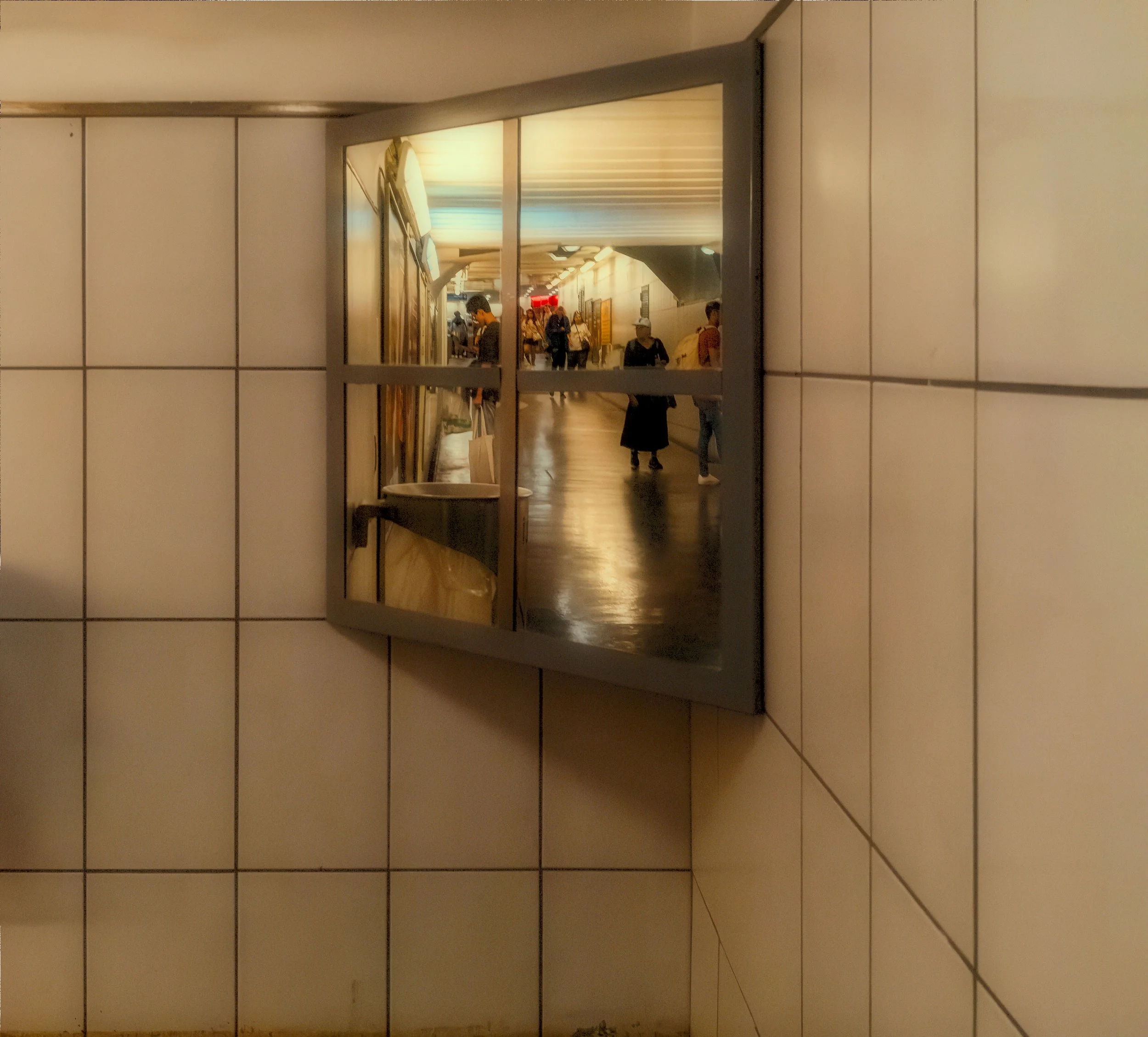 Interior corridor reflected in a wall mirror, showing people moving through a warm-lit passage framed by tiled walls.