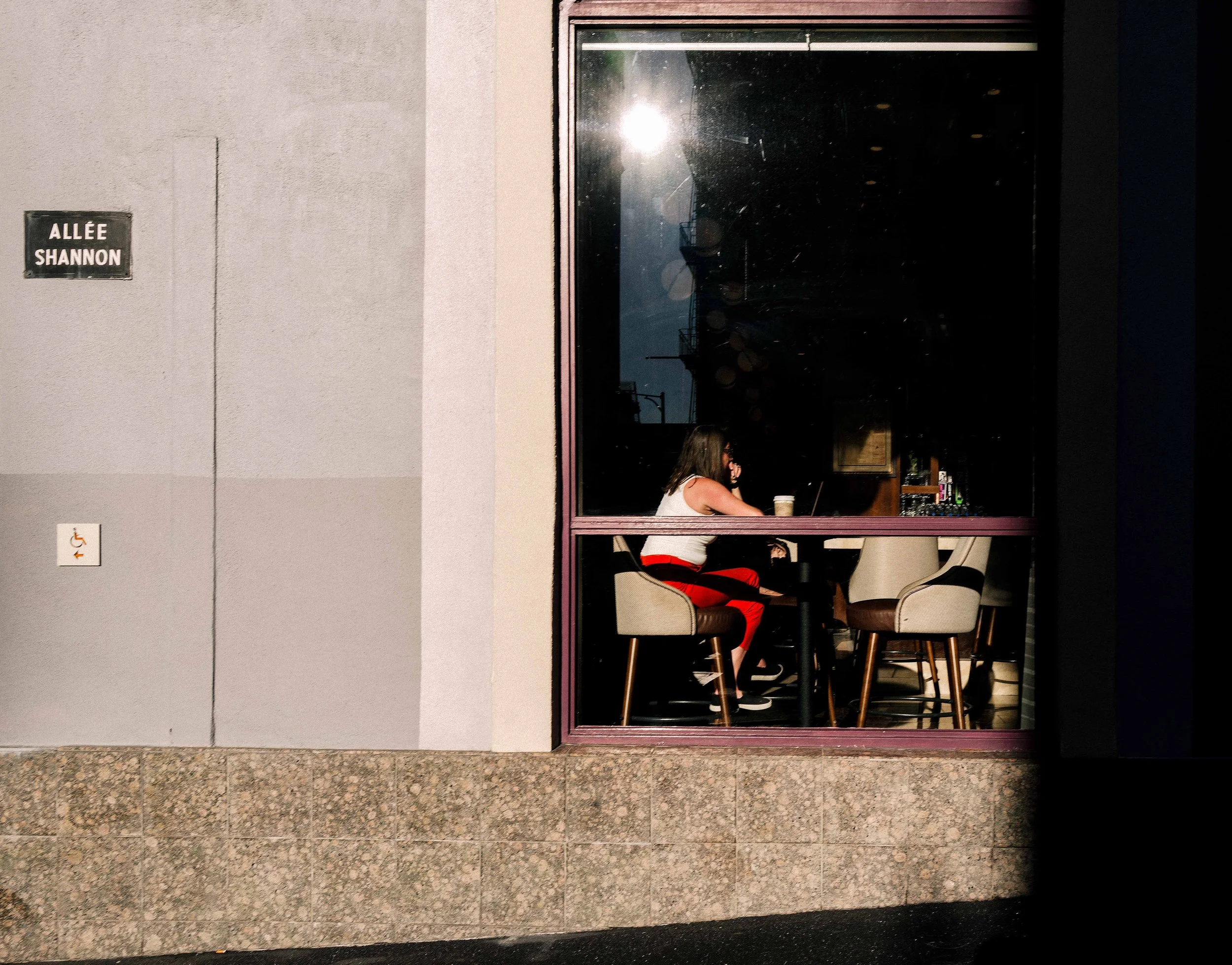 Woman seated alone inside a café or restaurant window on Allée Shannon, framed by strong sunlight, shadow, and a quiet interior.