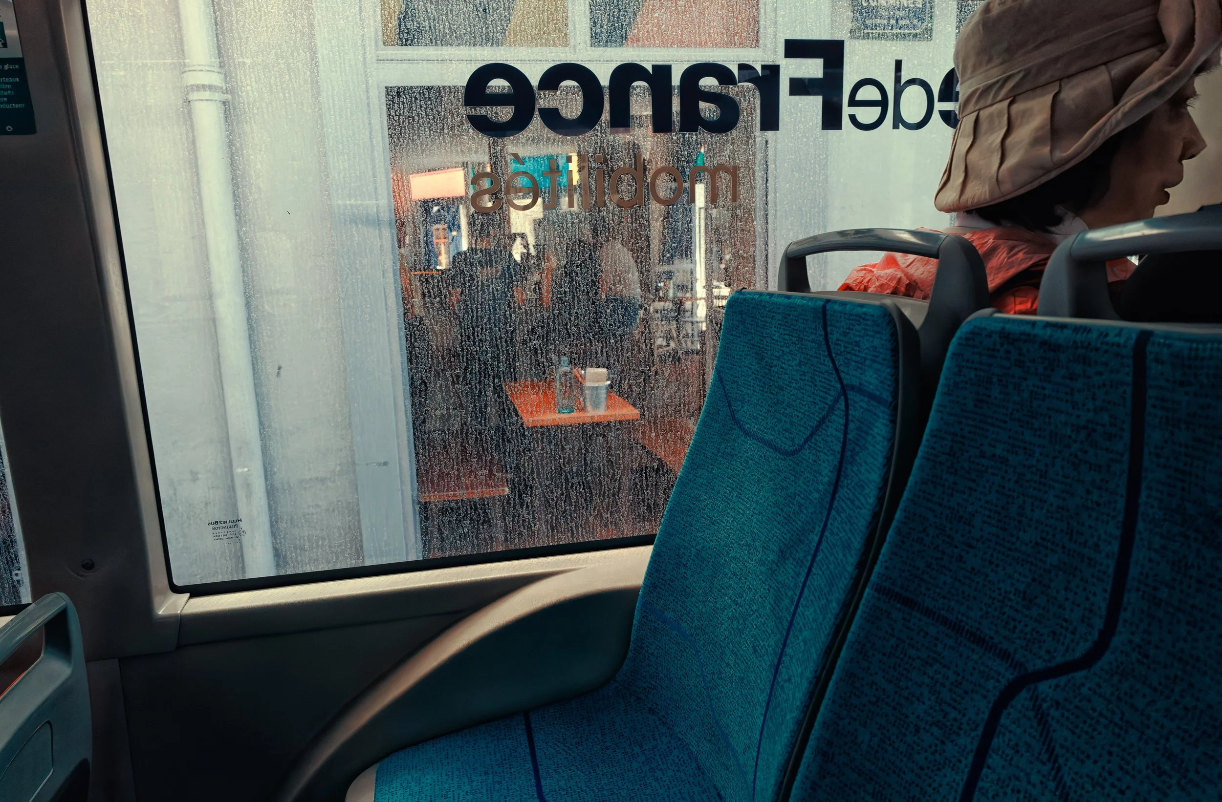 Half-Seen Passage: A passenger seen from behind sits on a blue-patterned bus seat beside a rain-speckled window, with blurred street and interior reflections visible through the glass.