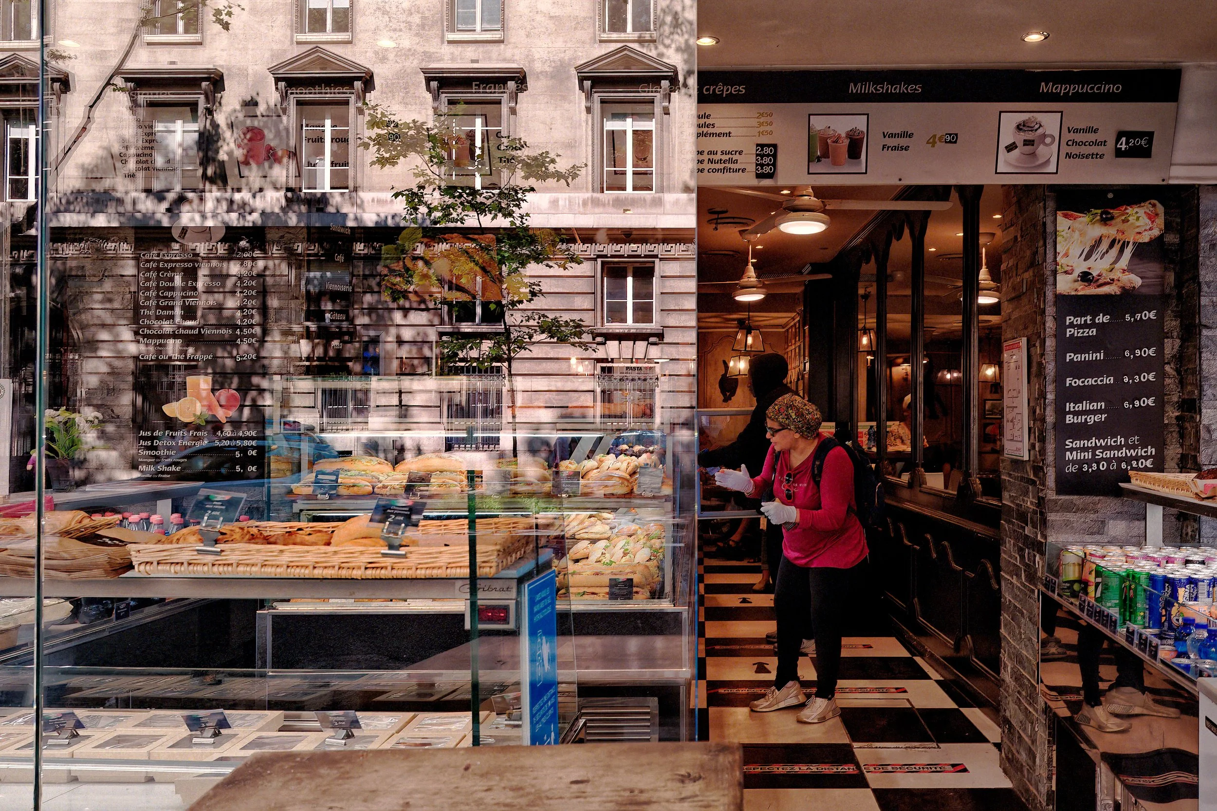 "Between Street and Café" (Paris)
Through the café window, the street and interior briefly occupy the same space. Stone façades settle over menus, bread displays, ceiling lights, and a worker moving through the room. Her exposed finger, pushing throu