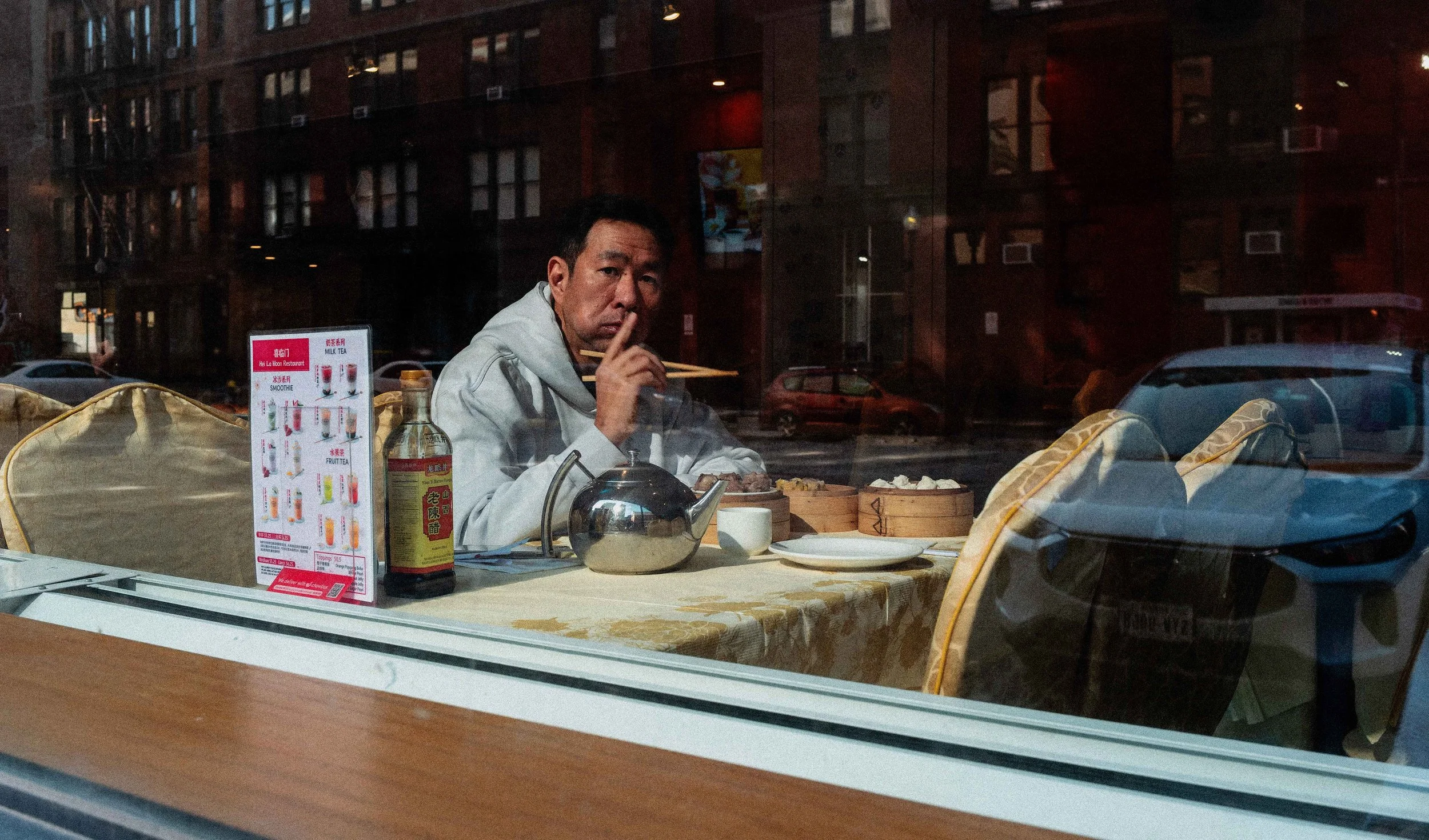 Man seated at a Chinatown restaurant table behind a window, with dim sum, tea, menu cards, and layered street reflections across the glass.