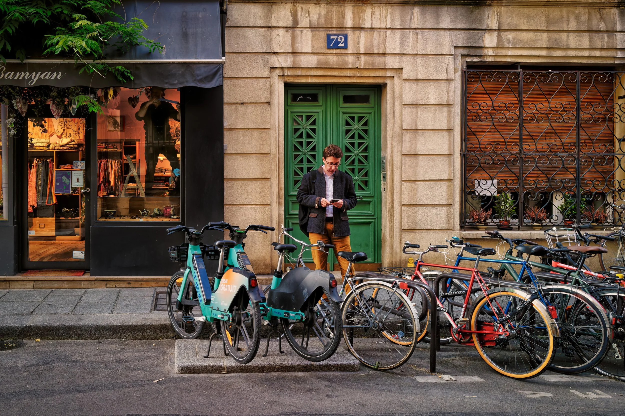 Number 72: A man in orange trousers checks his phone beside a green door, with parked bicycles in front and a shadowy figure visible in a warmly lit shop window.