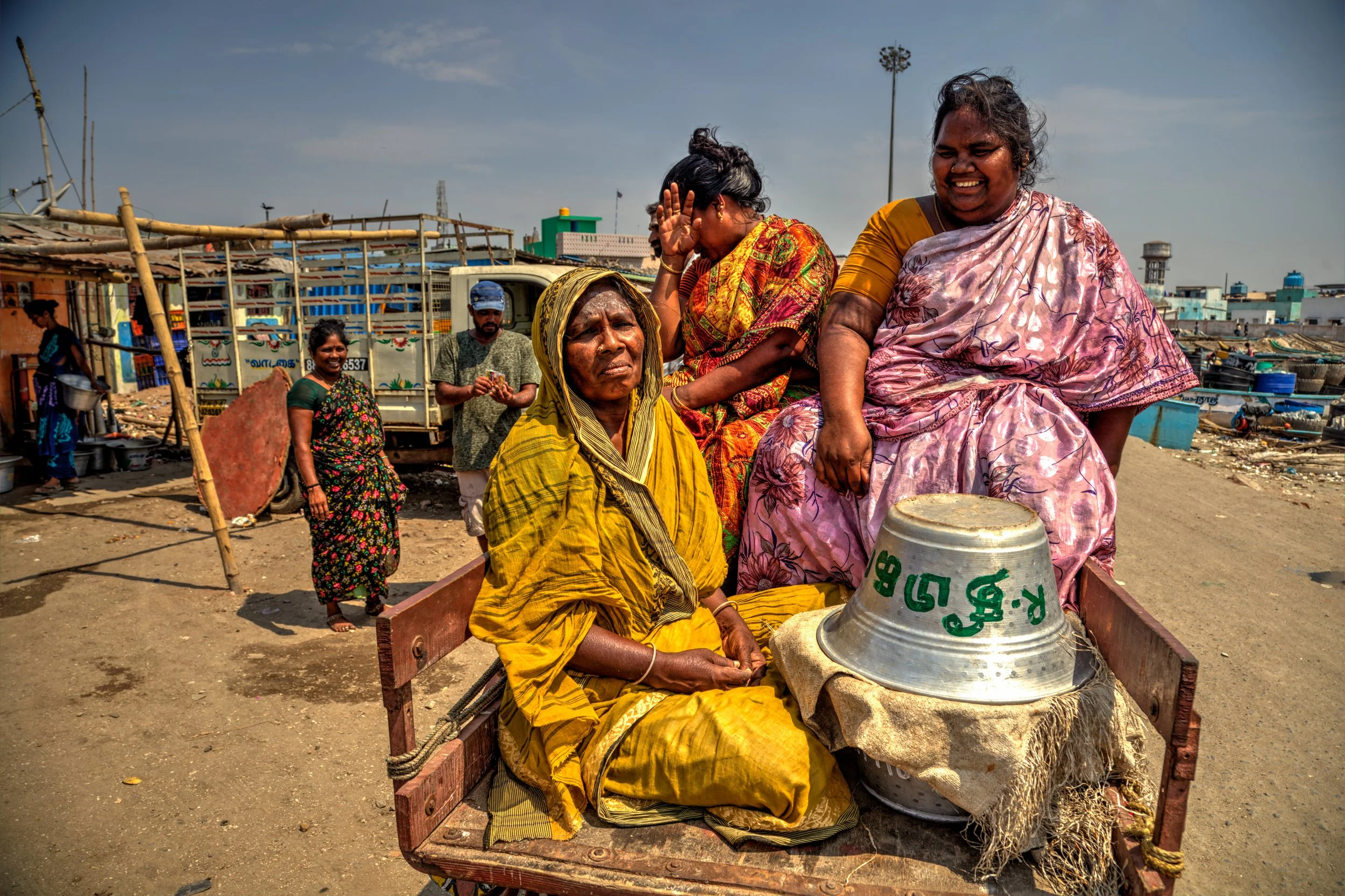 Women in colorful saris seated on a wooden cart near a harbor in India, with metal vessels, boats, workers, and bright midday light around them.