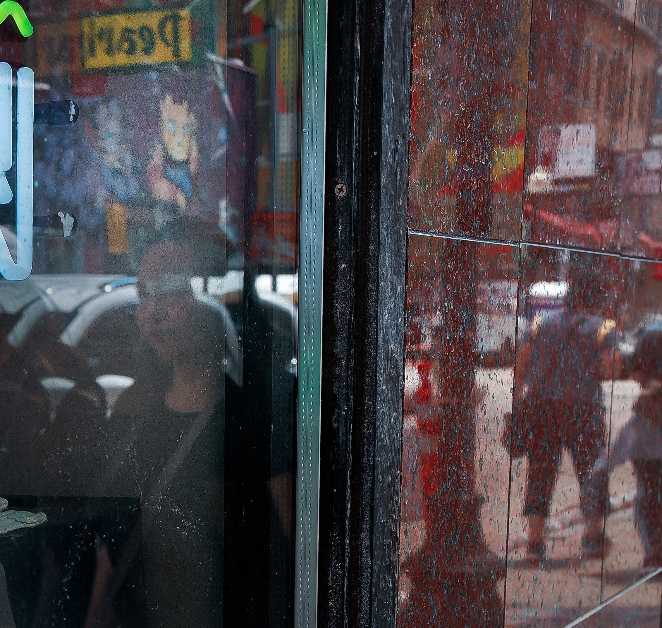 Shadowed figure seen through layered glass and reflections, with a red textured wall and blurred street details beside the window.
