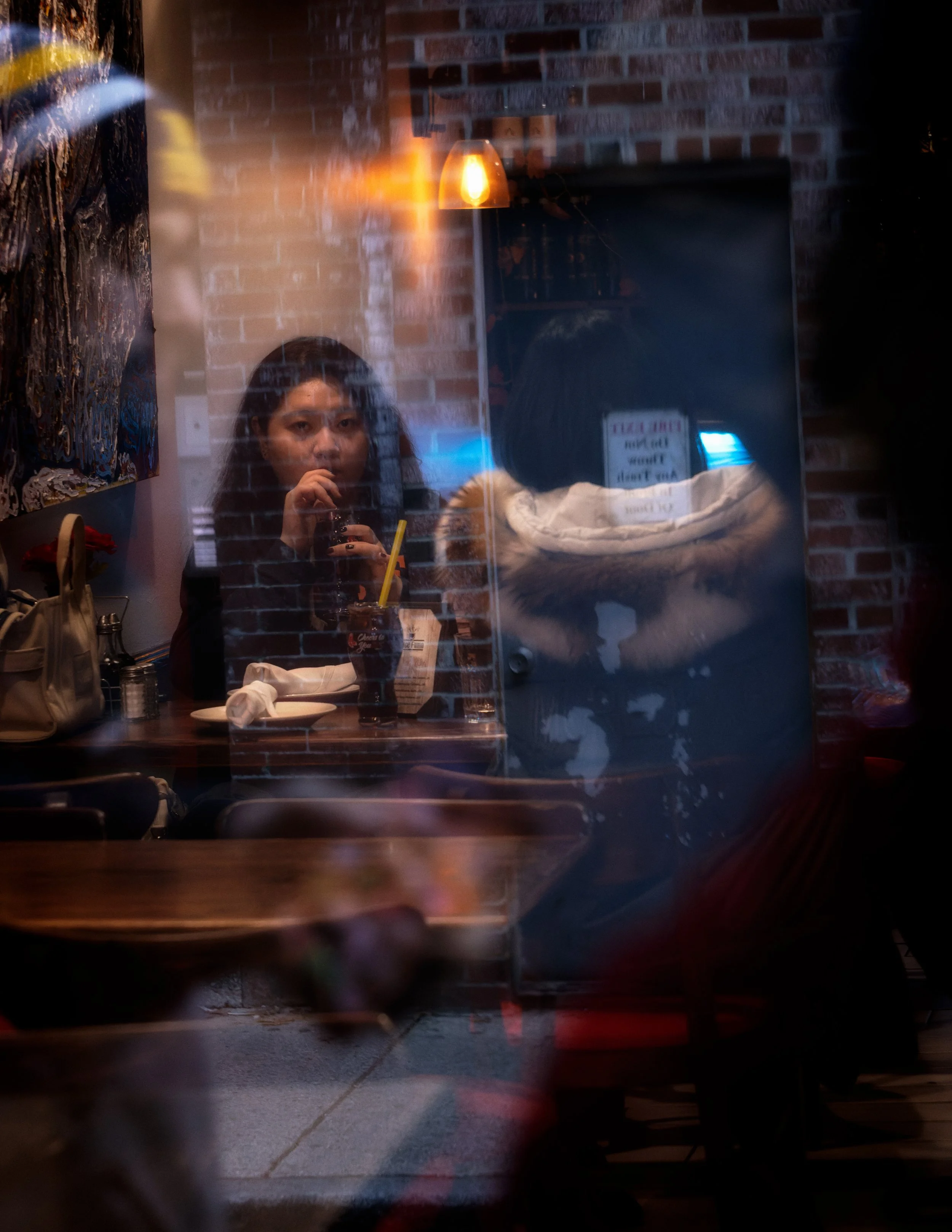 Woman seated at a restaurant table seen through layered window reflections, with drinks, brick walls, and another diner in a fur-trimmed coat.