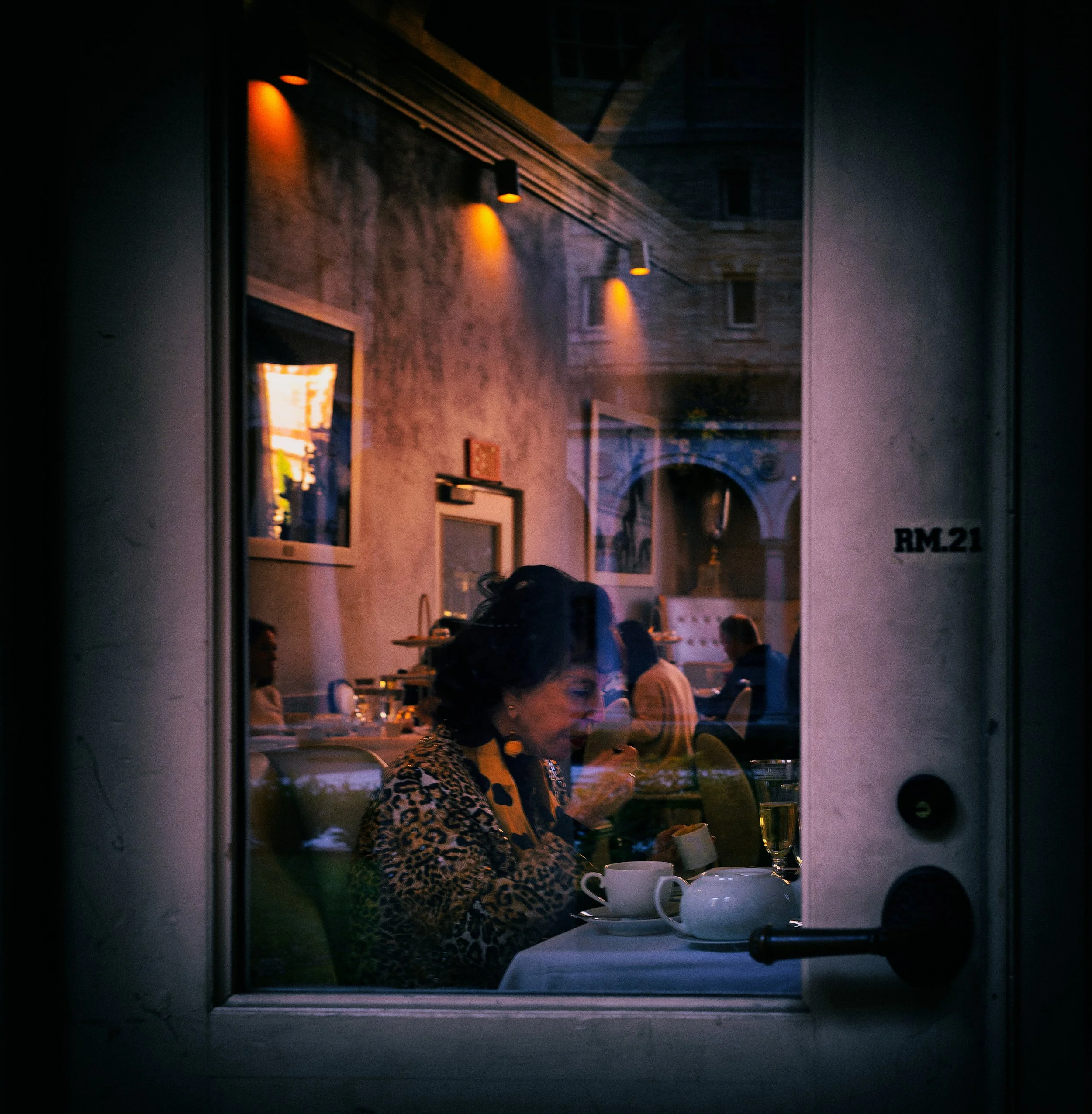 Woman seated inside a warmly lit restaurant seen through glass, layered with reflections, interior diners, tableware, and the number 21 on the doorway.