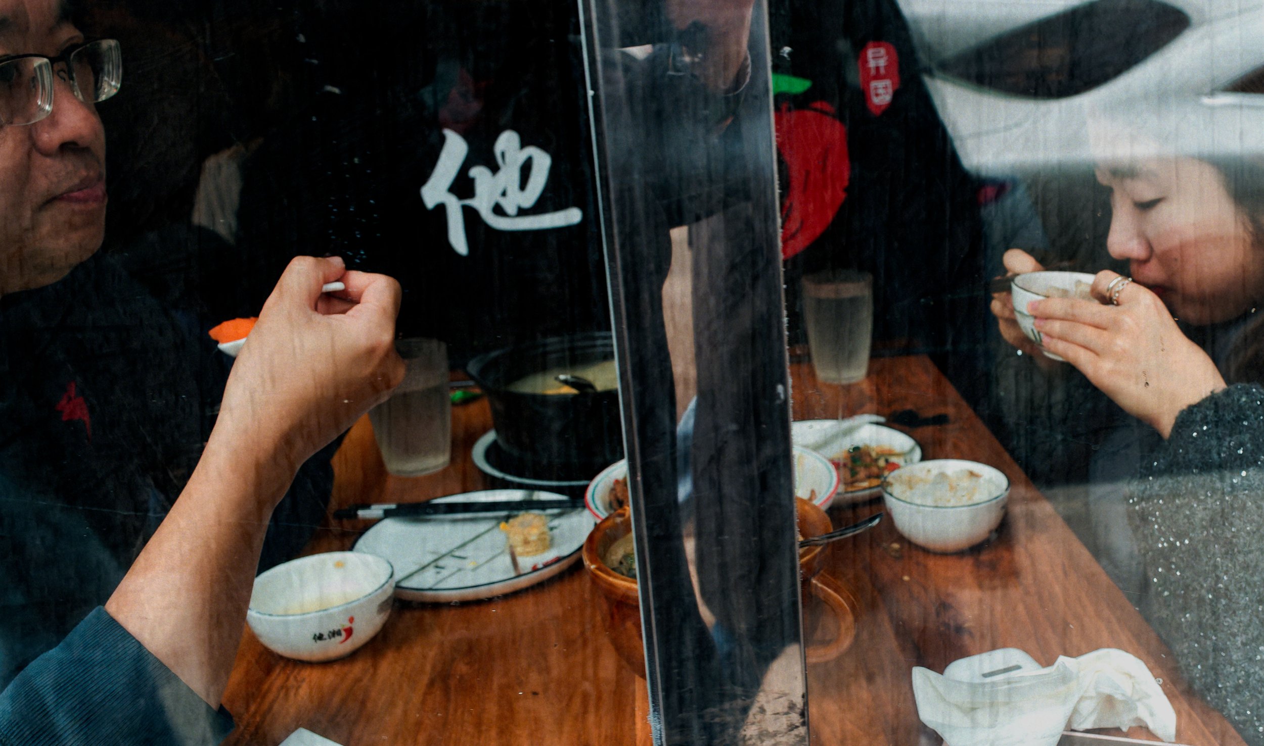Diners eating inside a Chinatown restaurant, seen through a window with layered reflections, bowls, hands, glass surfaces, and street details.
