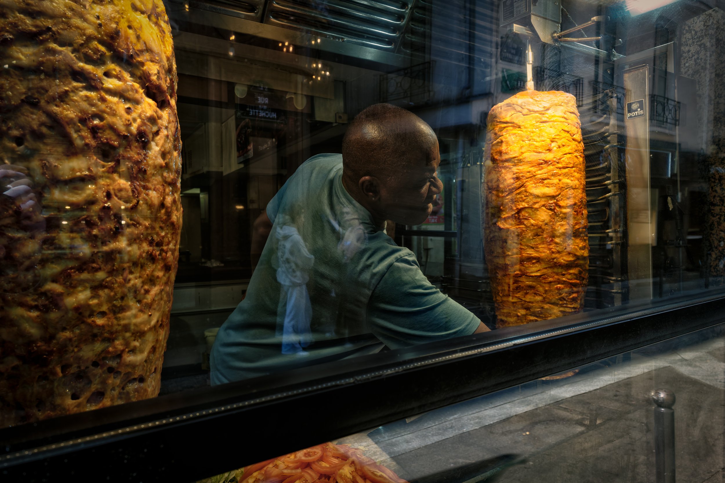 Between the Spits:
Behind the glass of a Paris kebab shop, a worker reaches between two towering vertical spits of roasting meat. Reflections of passing figures move across his shirt and the window, merging the activity of the street with the heat an
