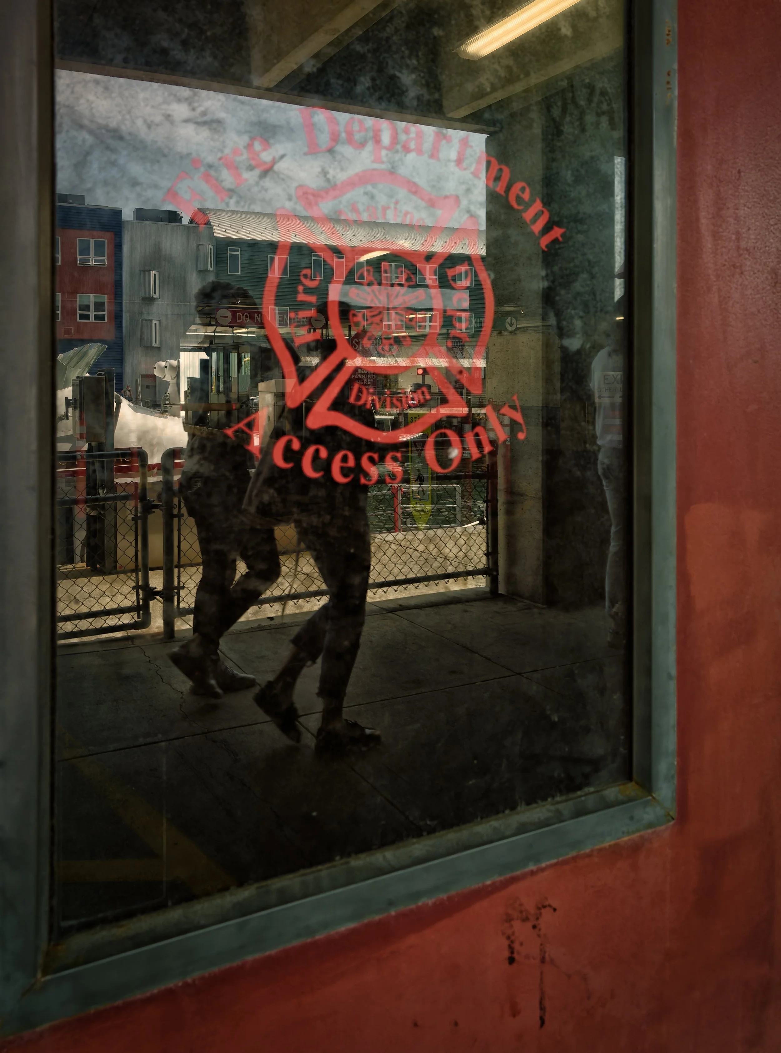 Pedestrian silhouette seen through layered glass reflections, with red “Access Only” signage, street barriers, and urban surfaces behind the window.