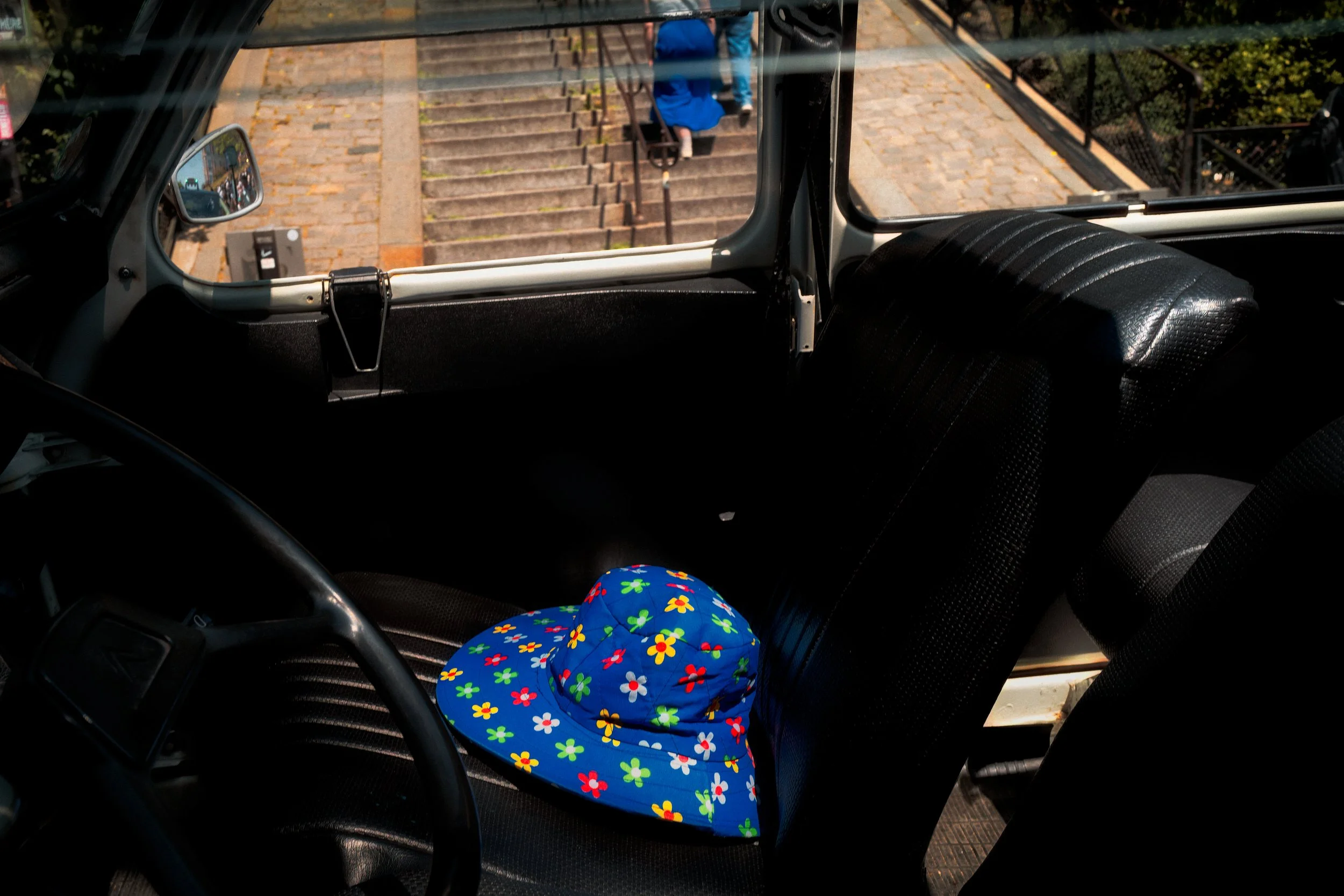 Blue Echo

In Paris, a bright floral hat rests on the seat of an otherwise empty vehicle while, beyond the window, a figure in blue climbs the steps outside. Separated by glass and distance, the two flashes of color quietly mirror one another, turnin