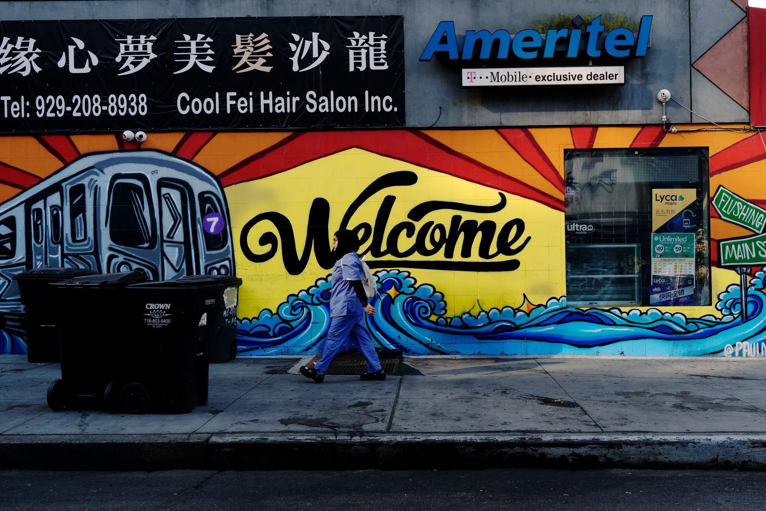 A woman in blue crosses a Flushing sidewalk beneath a vivid mural, her stride echoing the painted waves and the oversized word “Welcome.” Color, movement, and signage briefly align in an ordinary street moment.