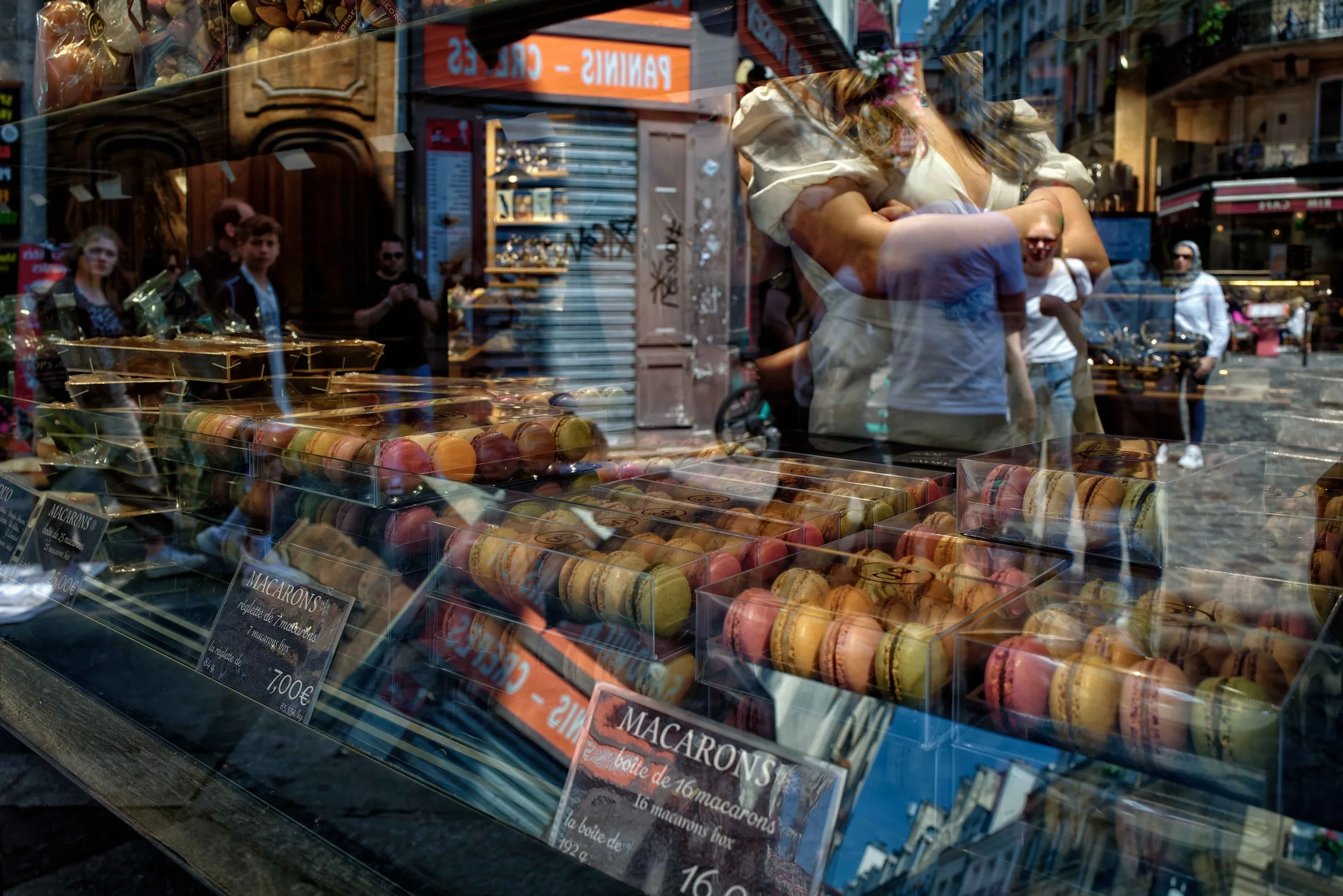 Layers of Sweetness:
Behind the glass, rows of macarons mingle with passing figures, folded arms, storefronts, and fragments of the street. What begins as a display of color and sweetness becomes something more layered—a moment in which interior and 