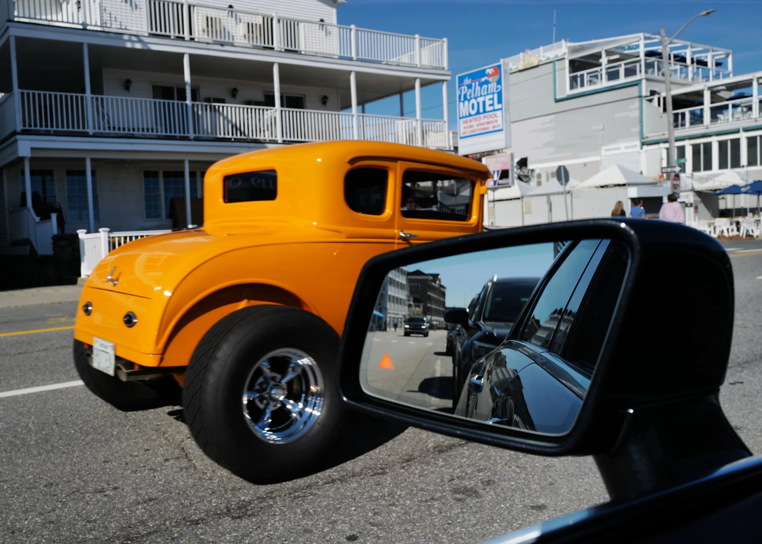 Orange vintage car passing beside a beach motel, with the street and vehicles reflected in a car side mirror.