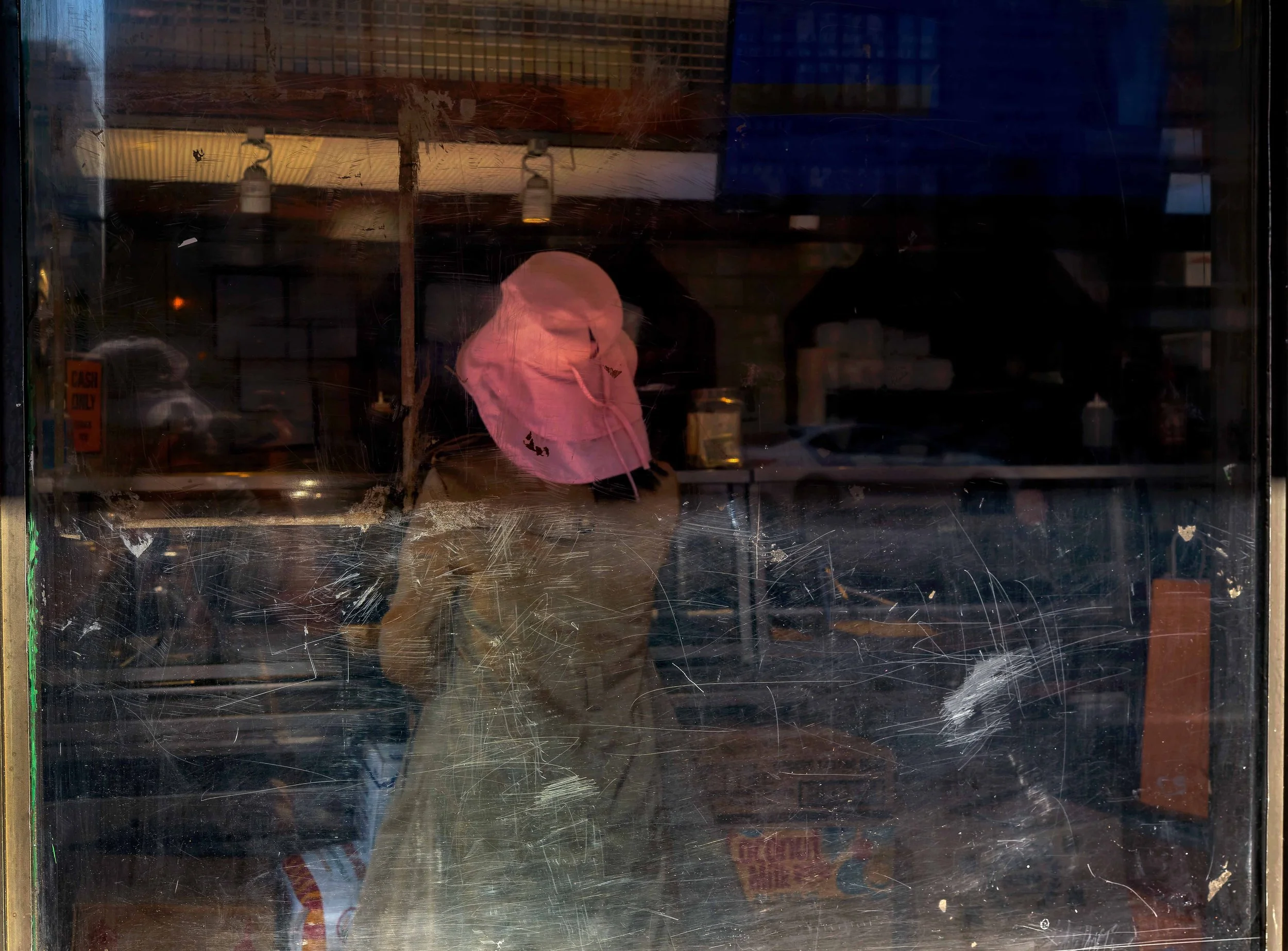 Figure wearing a pink hat seen through scratched and reflective glass, layered with dark interior shadows, street reflections, and window surface marks.
