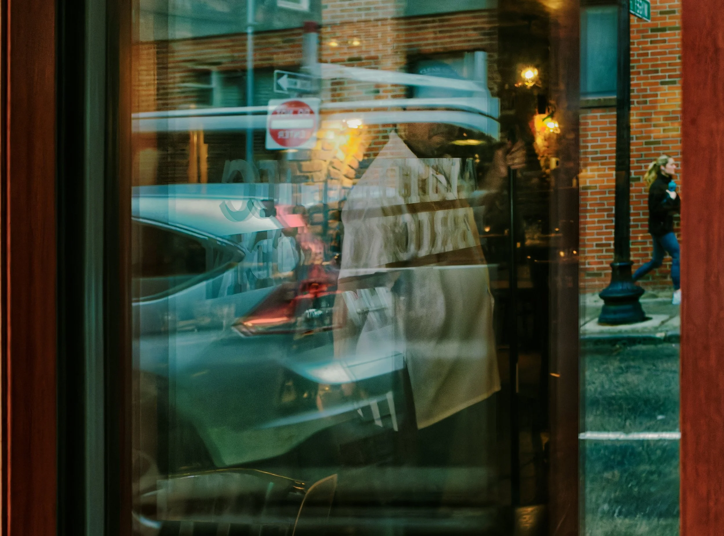 Layered window reflection showing an interior figure overlapping with a parked car, street signs, brick buildings, and a pedestrian outside.