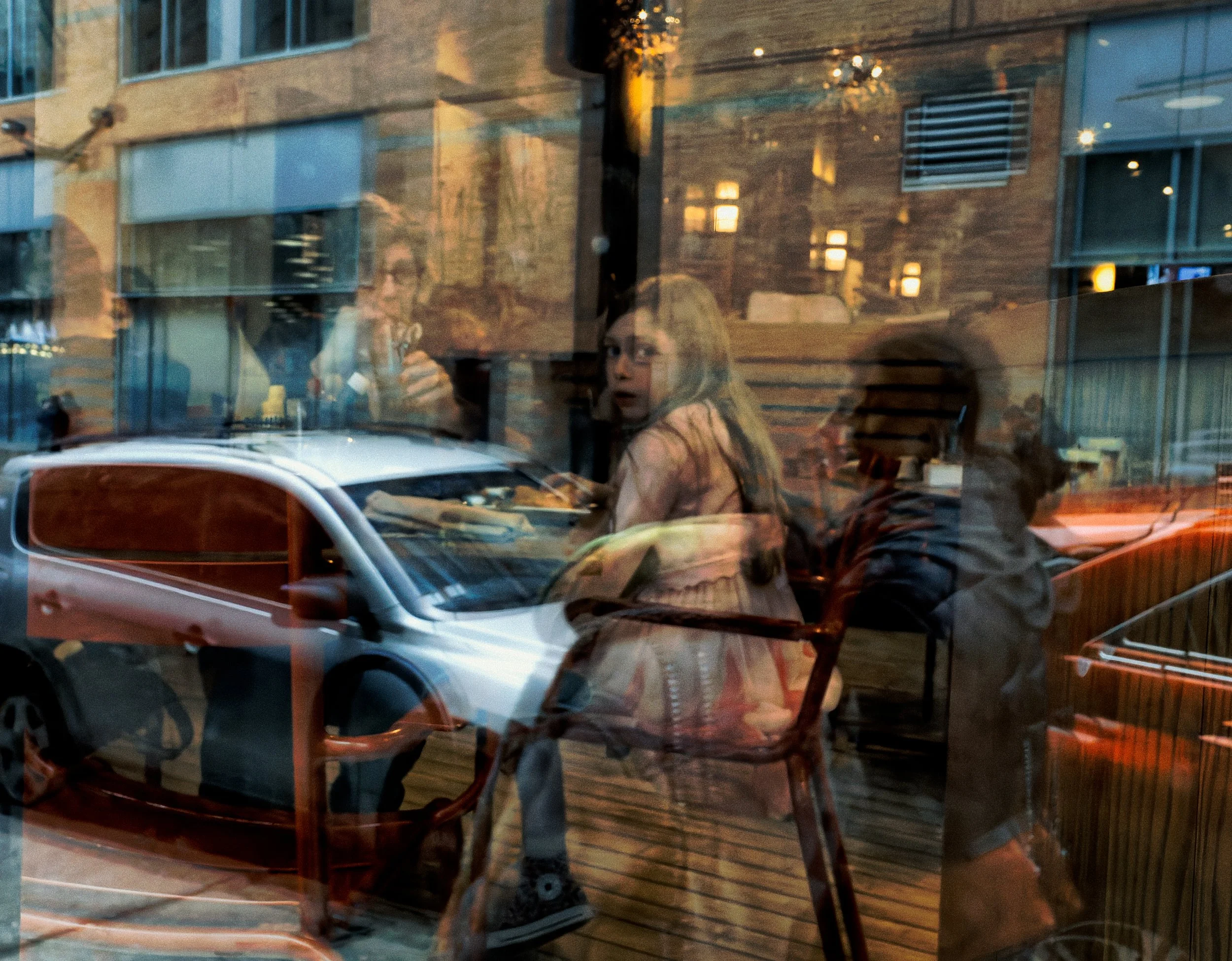 People seen through a restaurant or café window, layered with reflections of a white car, city buildings, warm interior lights, and street activity.