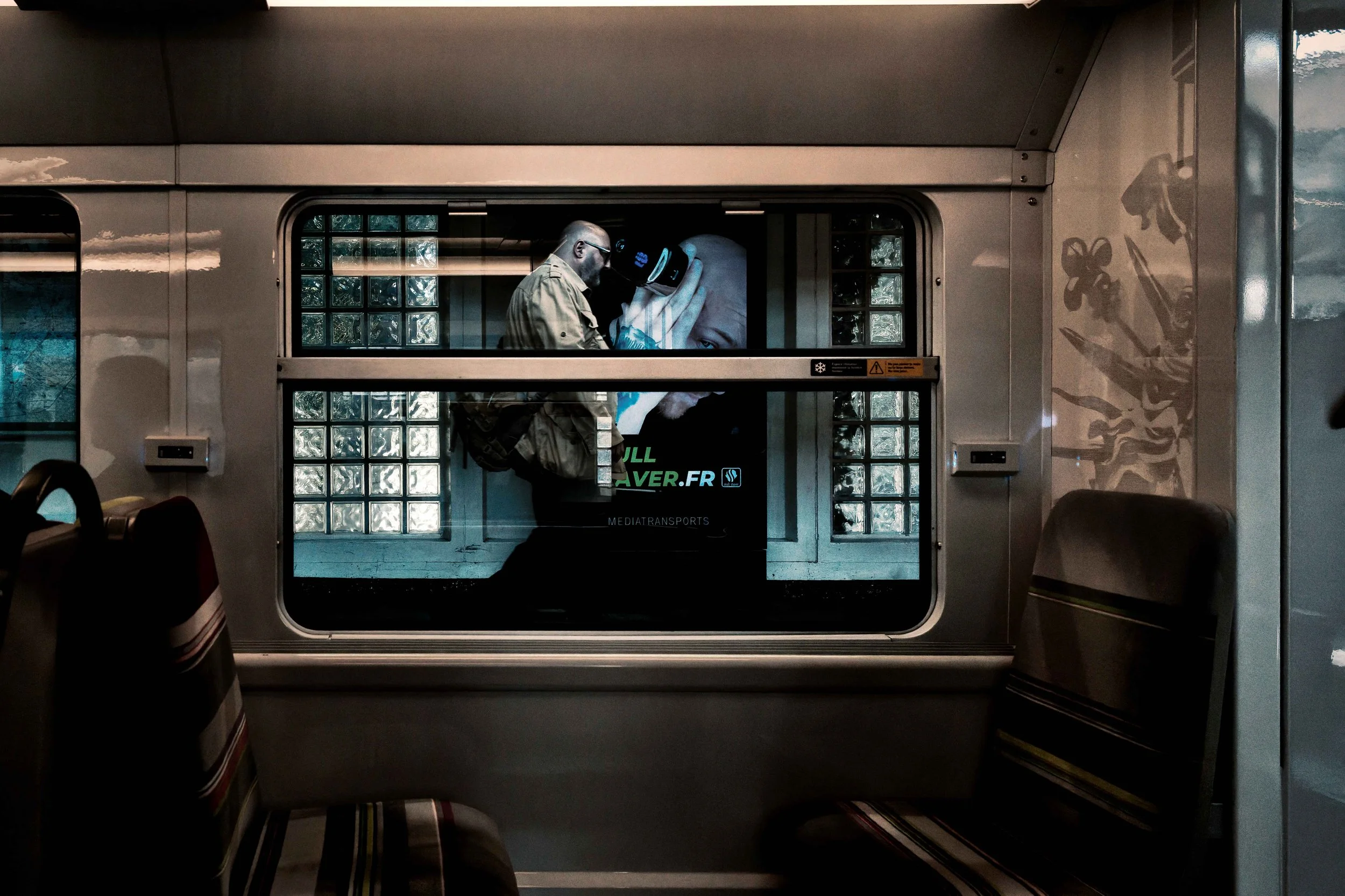 Passing Resemblance:
Inside a dim Paris Métro carriage, a passing man briefly aligns with an advertisement for a head shaver visible through the window. His closely shaved head mirrors the commercial figure behind him, while the enlarged hand and gro