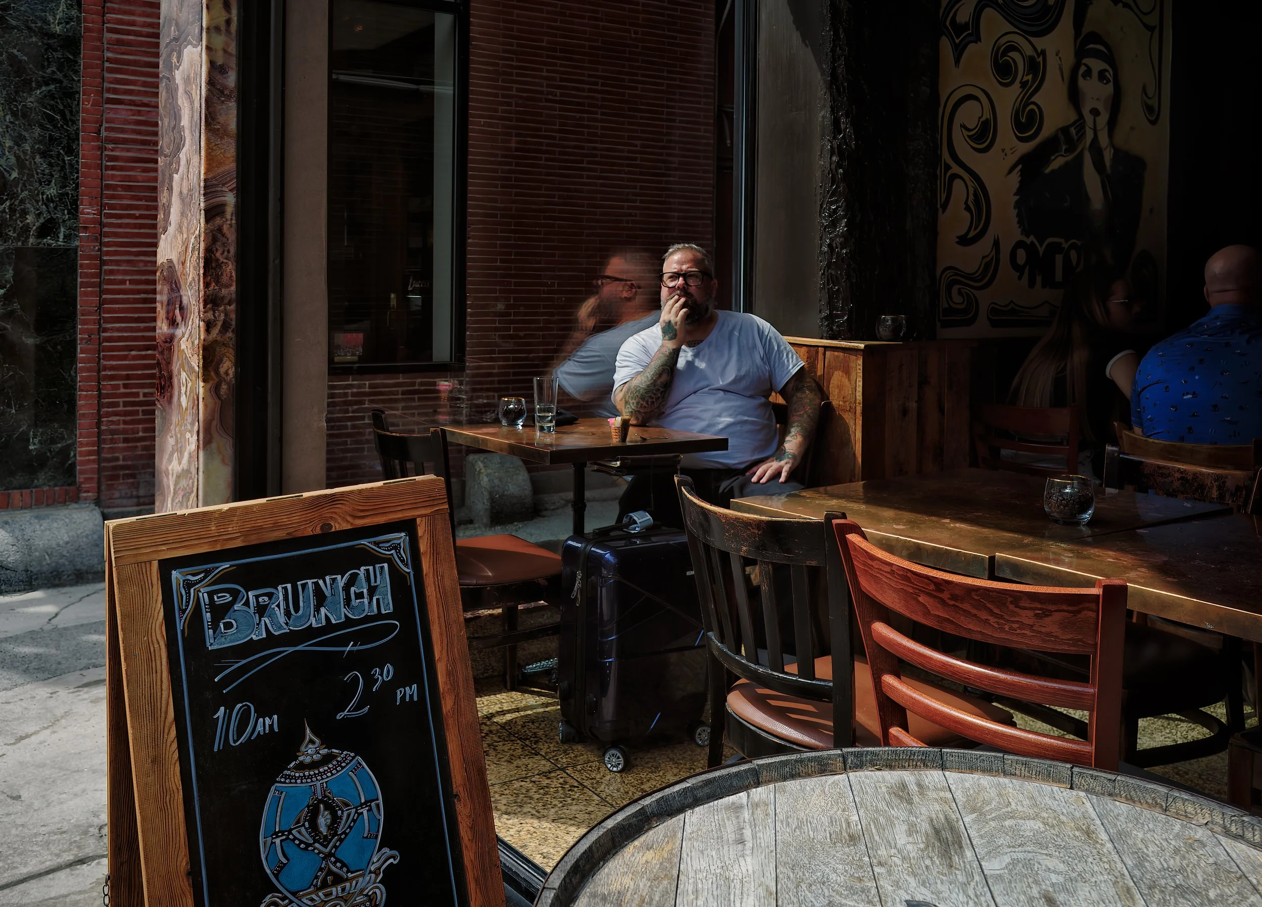 Man seated at a café table inside a dim interior, layered with window reflections, a brunch sign, empty chairs, and street details outside.