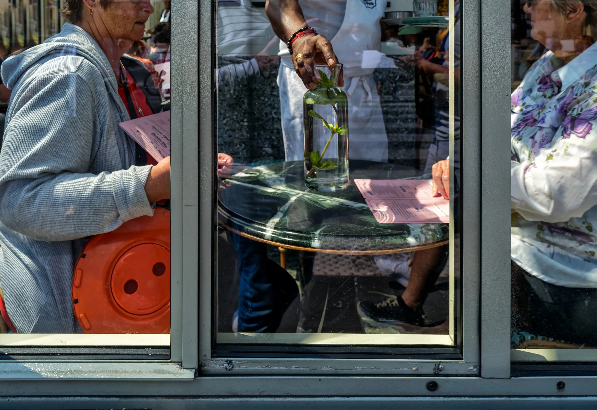 People seated around a café table in Paris seen through window frames, with a glass bottle, menus, hands, and layered reflections across the scene.